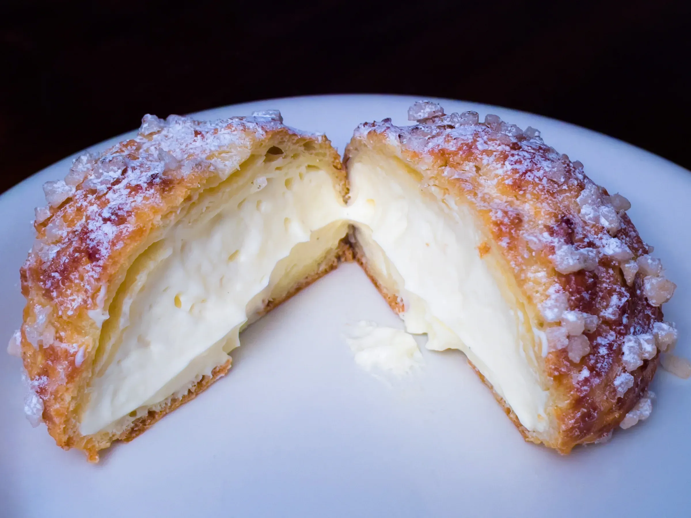 Large choux pastry split open on a white plate — thick vanilla cream filling, pearl sugar and powdered sugar on the golden crust