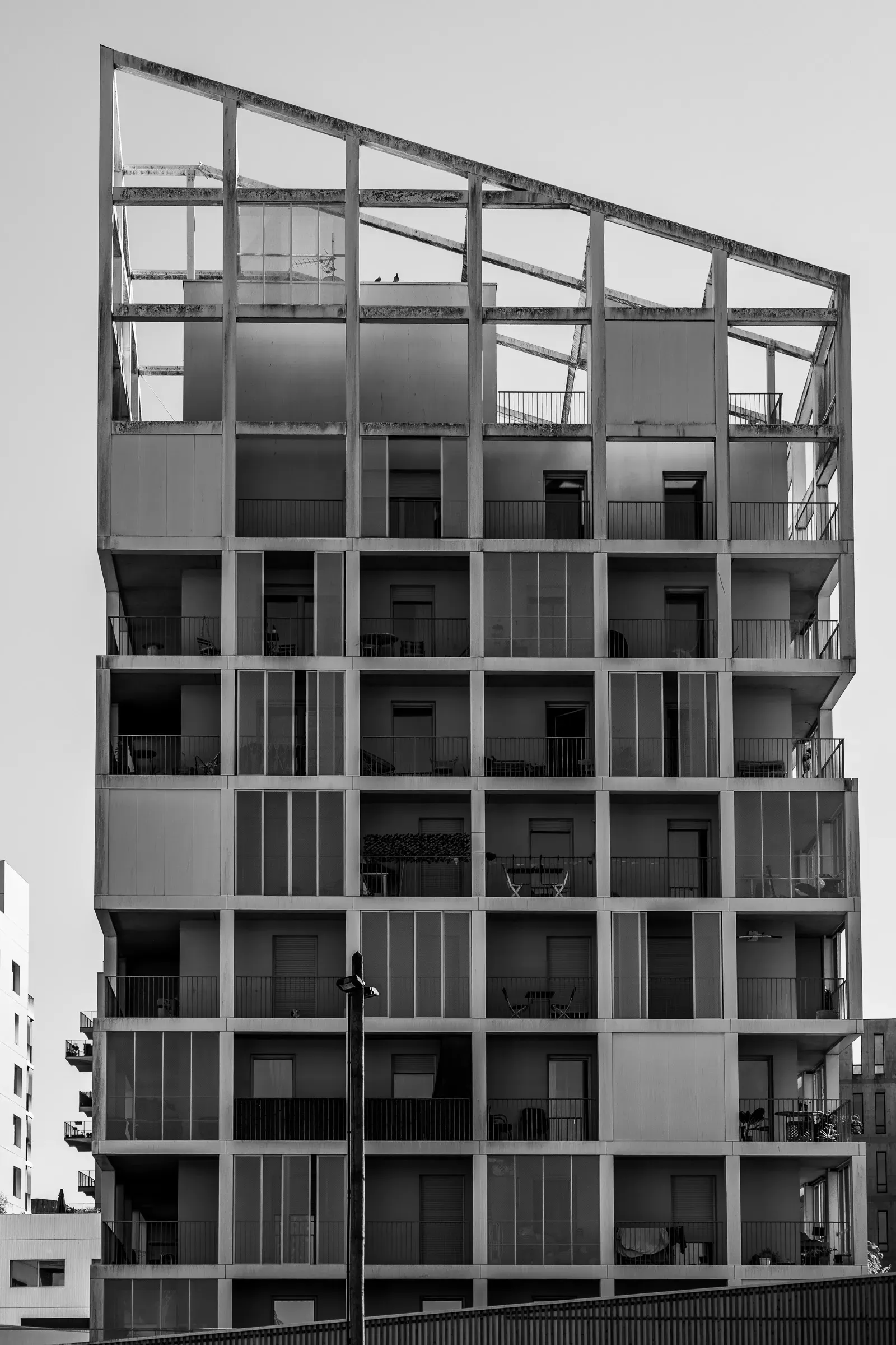 Modern residential building with a strict grid of balconies and horizontal panels, the top floor an open steel skeleton, against a pale sky