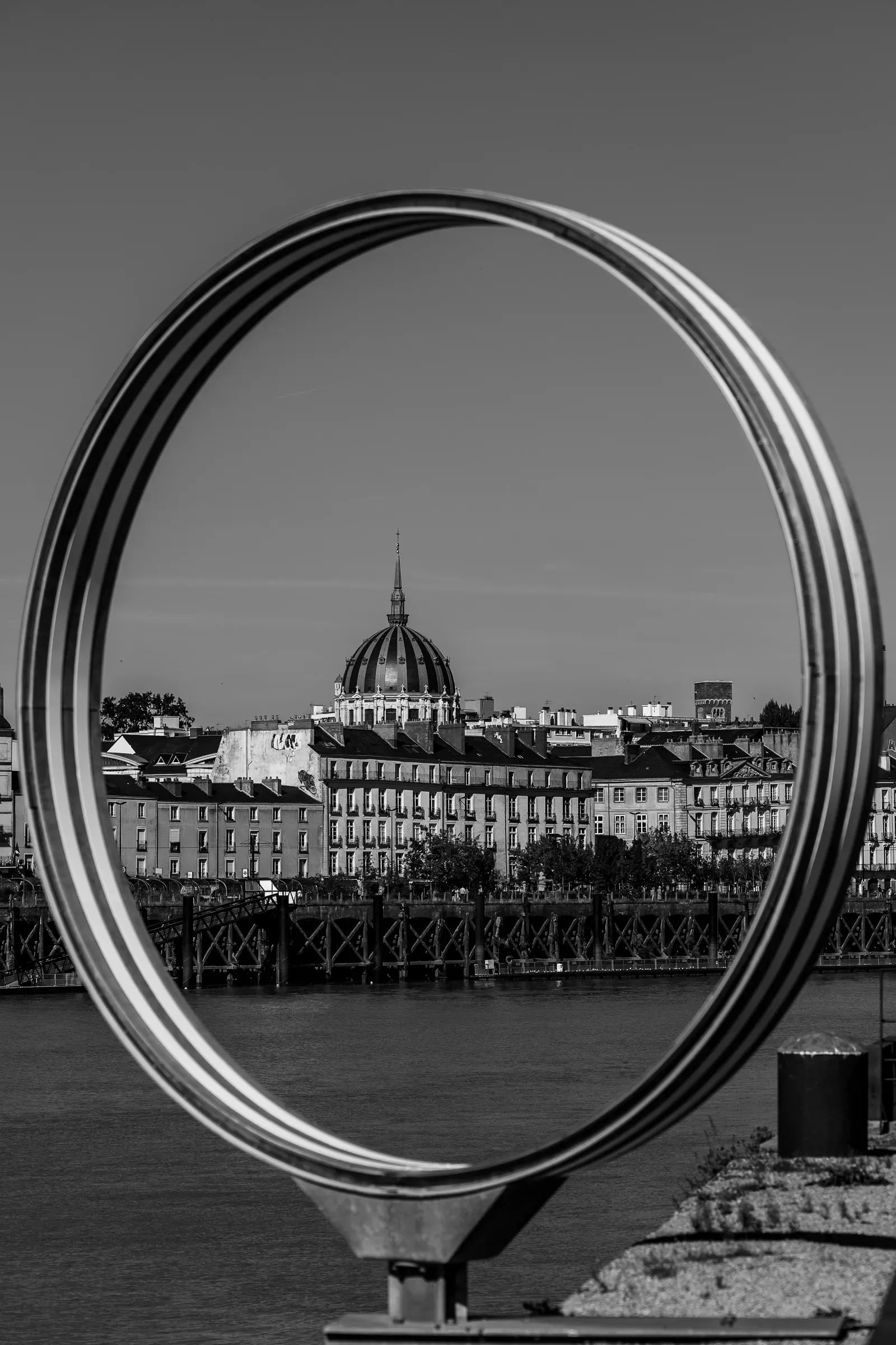 A circular polished-steel sculpture frames a domed building across a river, the city skyline and a bridge visible through the ring
