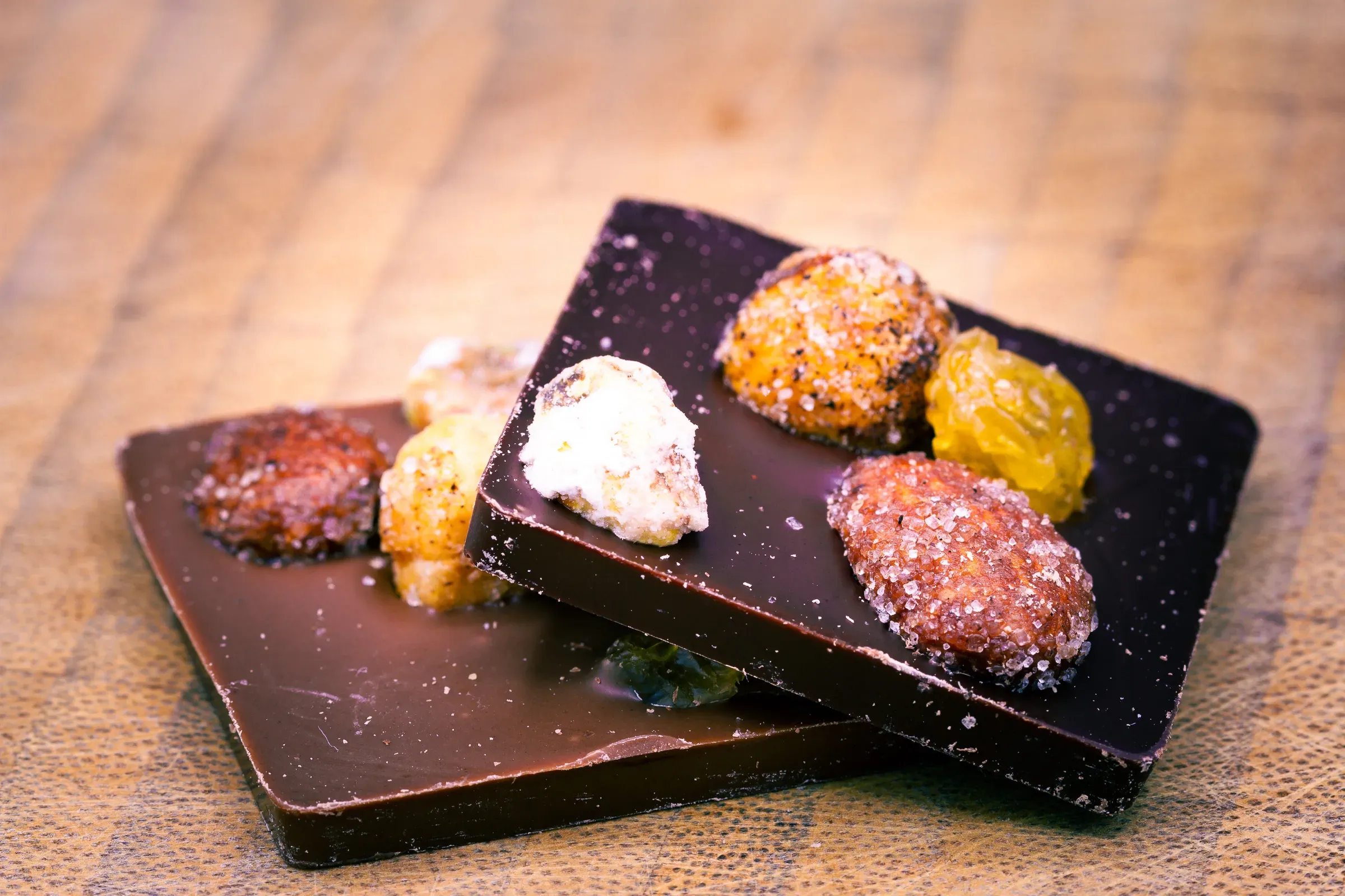 Two dark chocolate bark squares on a wooden surface, topped with candied and dried fruit pieces, stacked at an angle