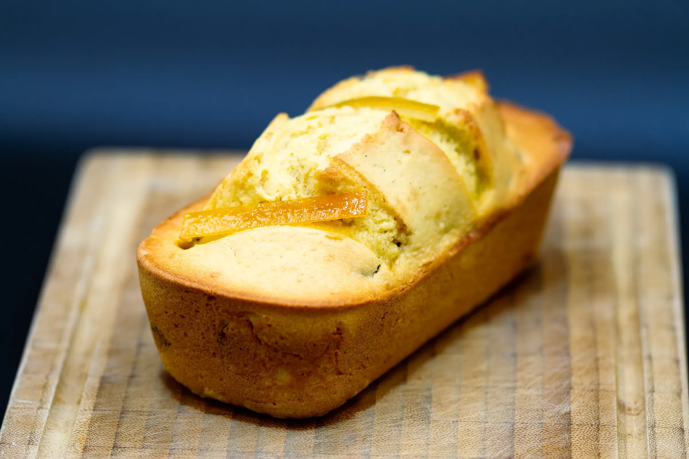 Clémentine loaf cake on a wooden board against a dark blue background — golden crust split along the top, fruit baked inside