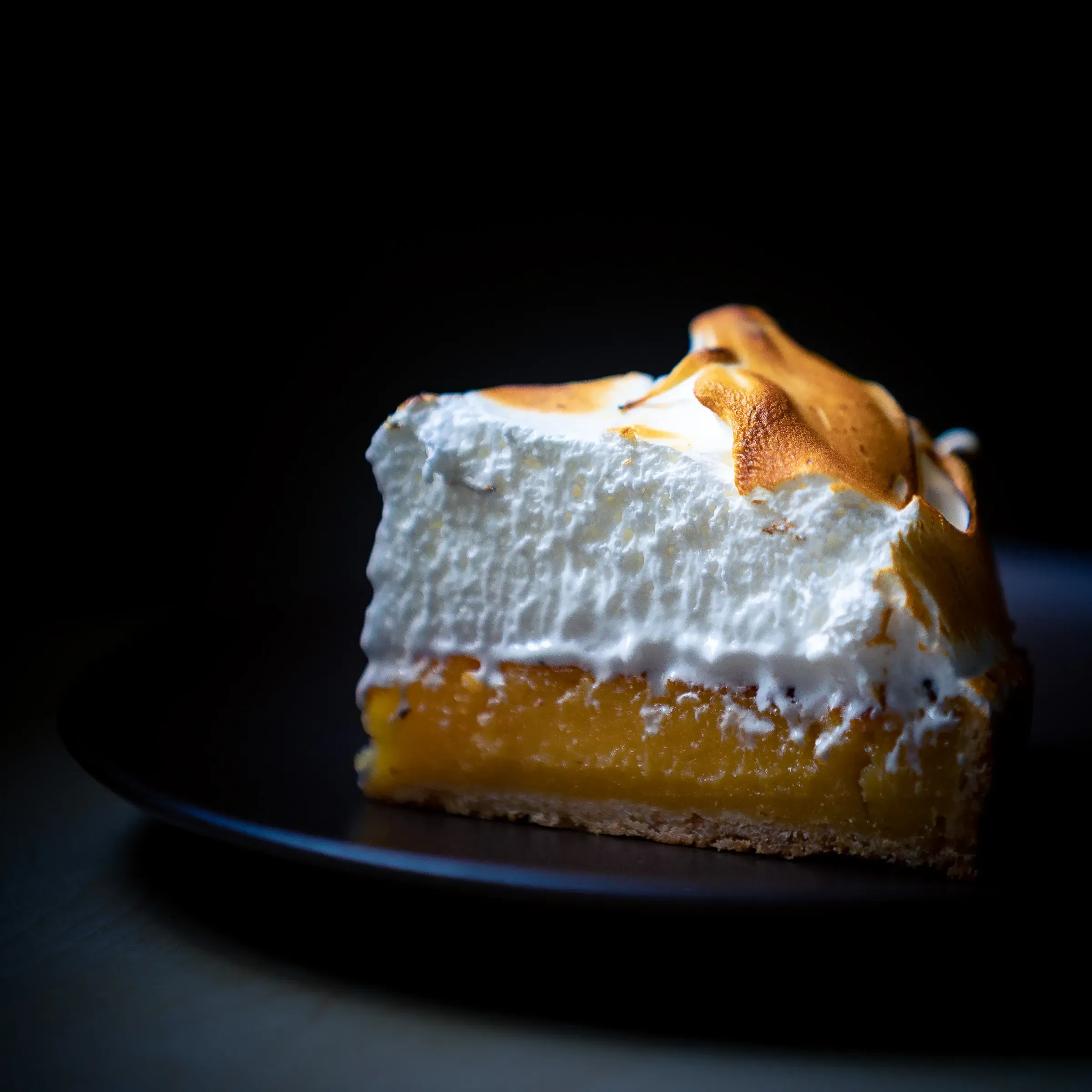 Slice of lemon meringue tart on a dark plate — thick torched meringue above bright yellow lemon curd, pastry base, black background