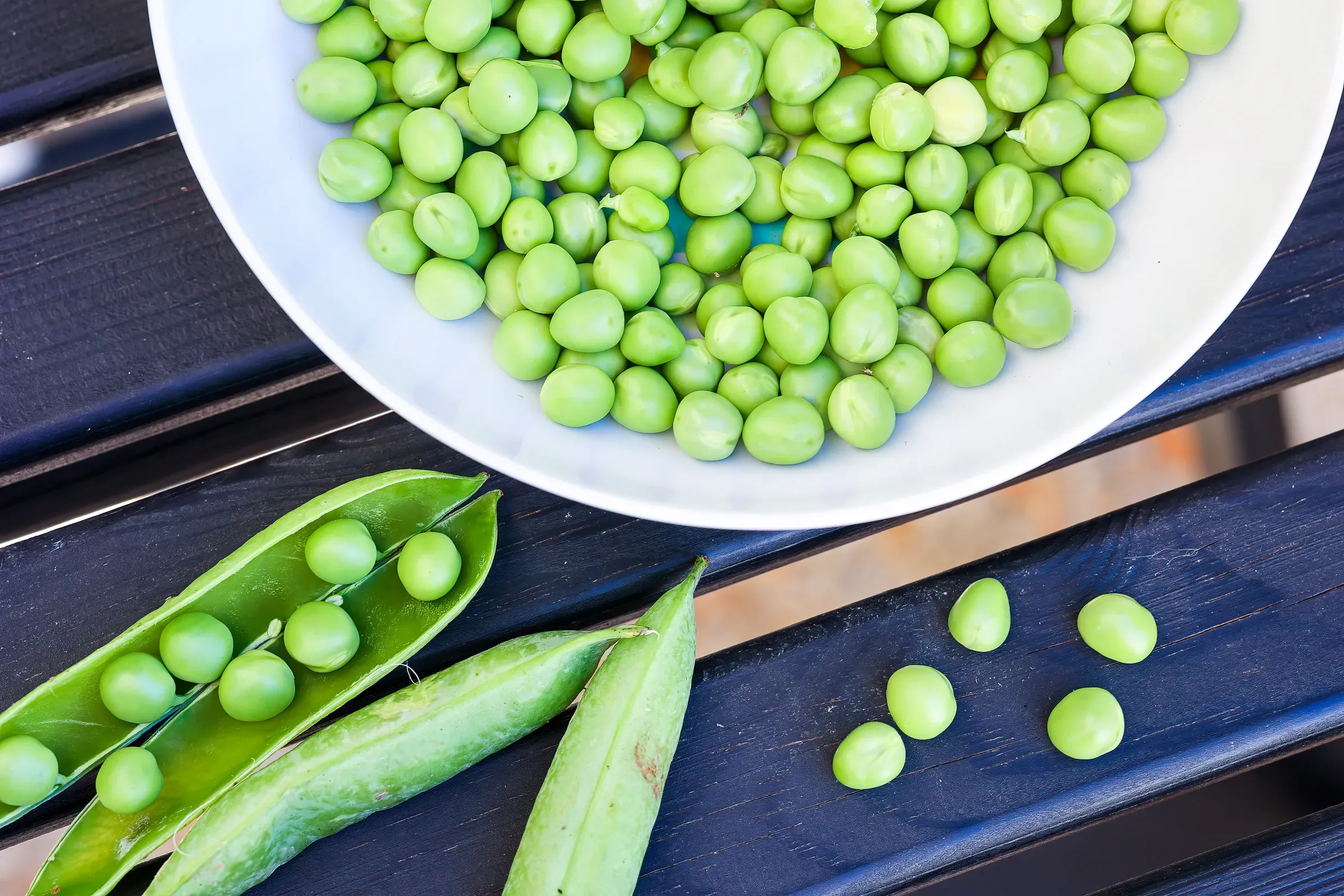 Overhead of a white bowl of freshly shelled peas on a dark slatted surface, two open pea pods laid beside it