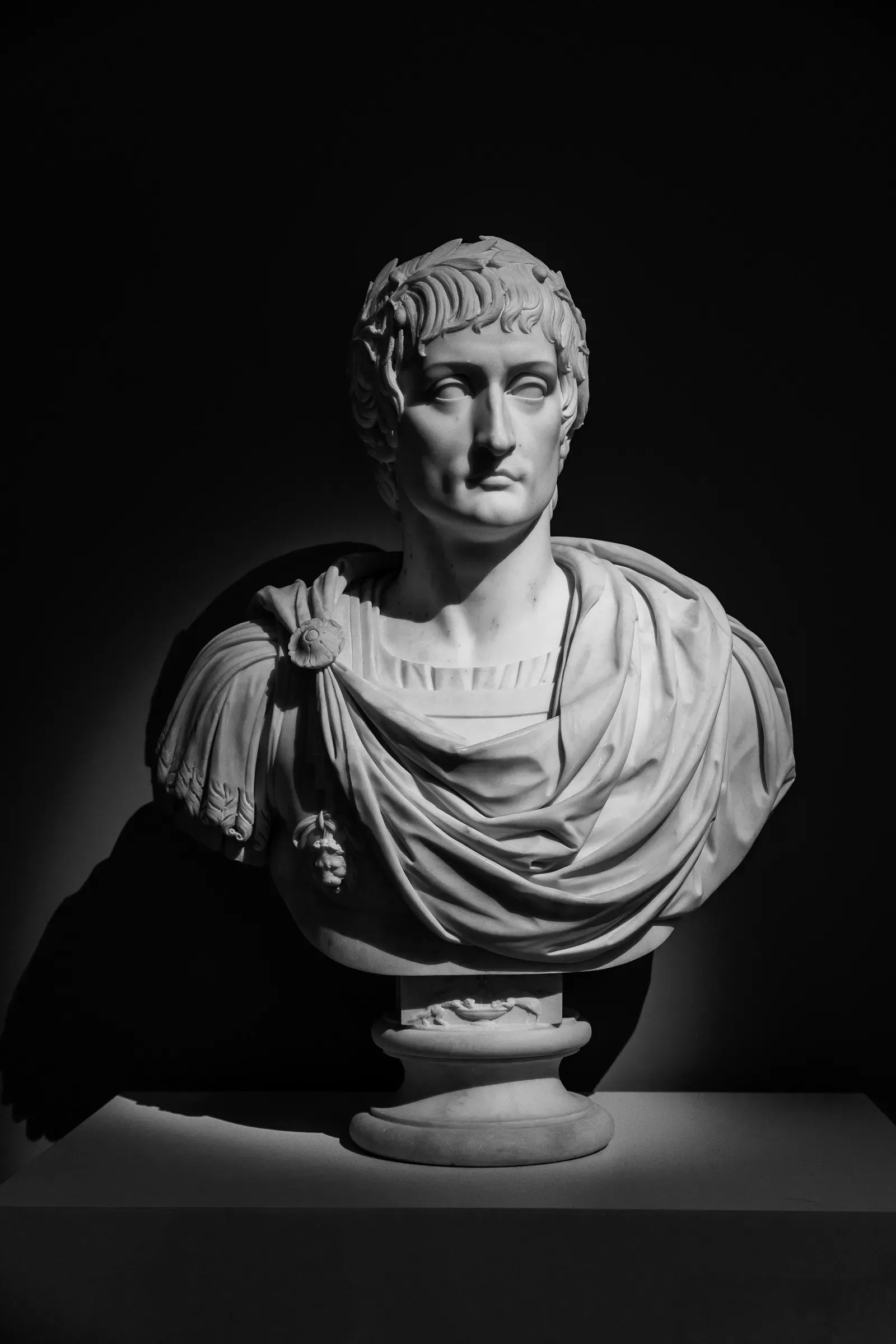 White marble Roman-style bust with laurel wreath and draped toga, lit dramatically against a pure black background, strong chiaroscuro
