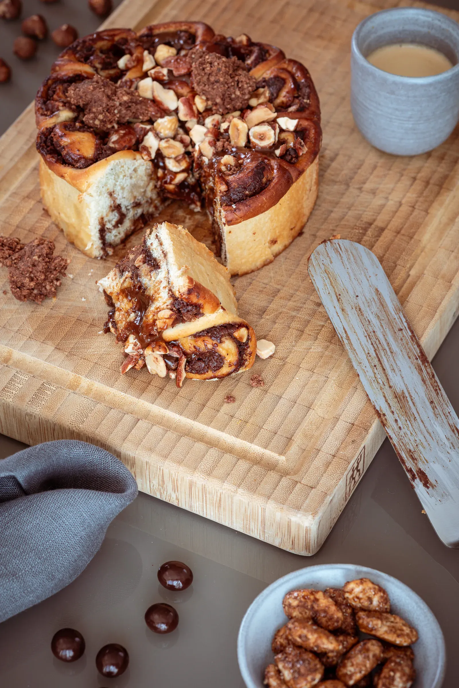 Chocolate hazelnut swirl bun cake on a wooden chopping board, a slice removed, cup of coffee and bowl of hazelnuts beside it