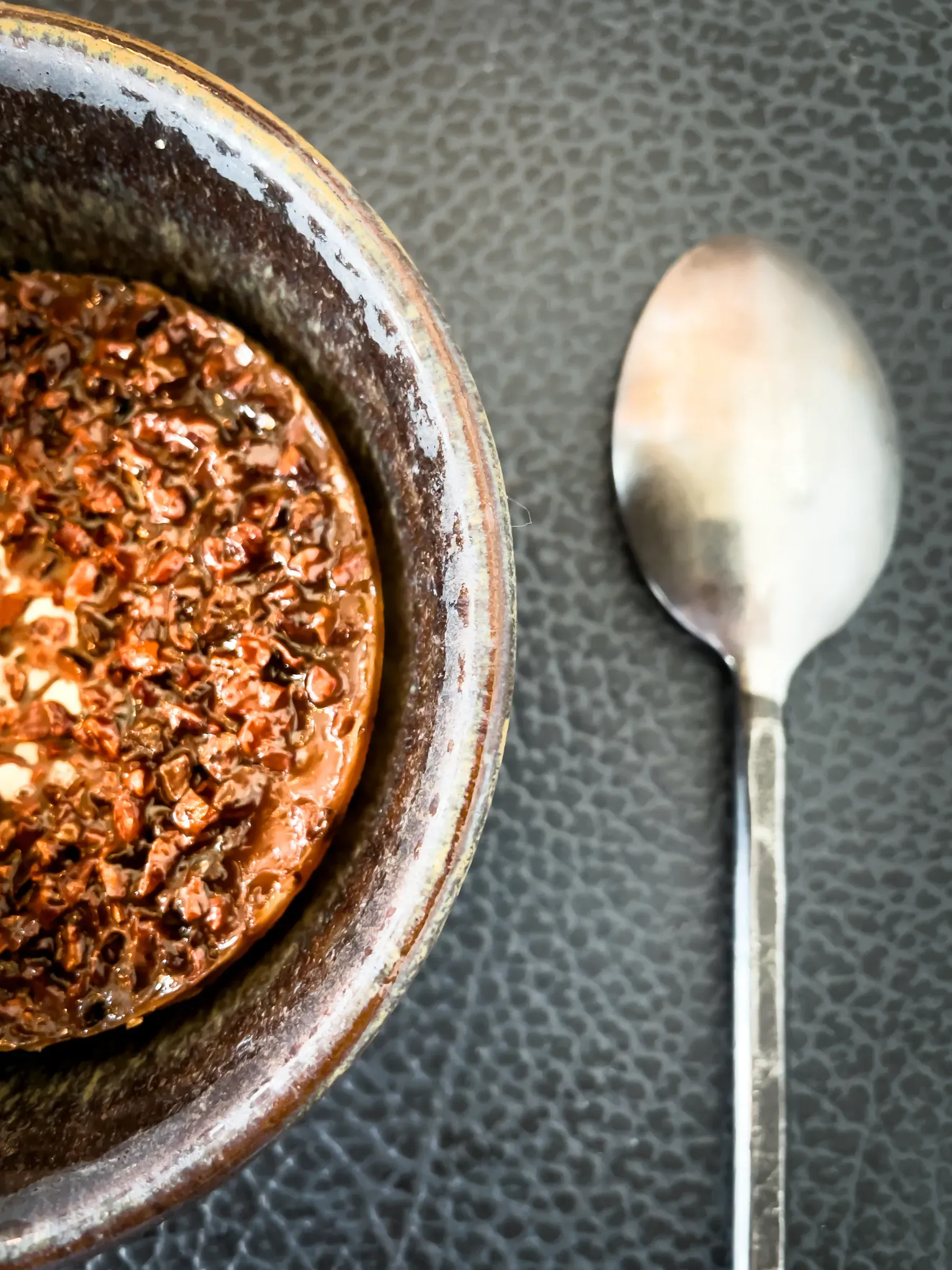 Dark stoneware bowl of chocolate pot de crème with cacao nibs on the surface, a silver spoon beside it on a dark textured background