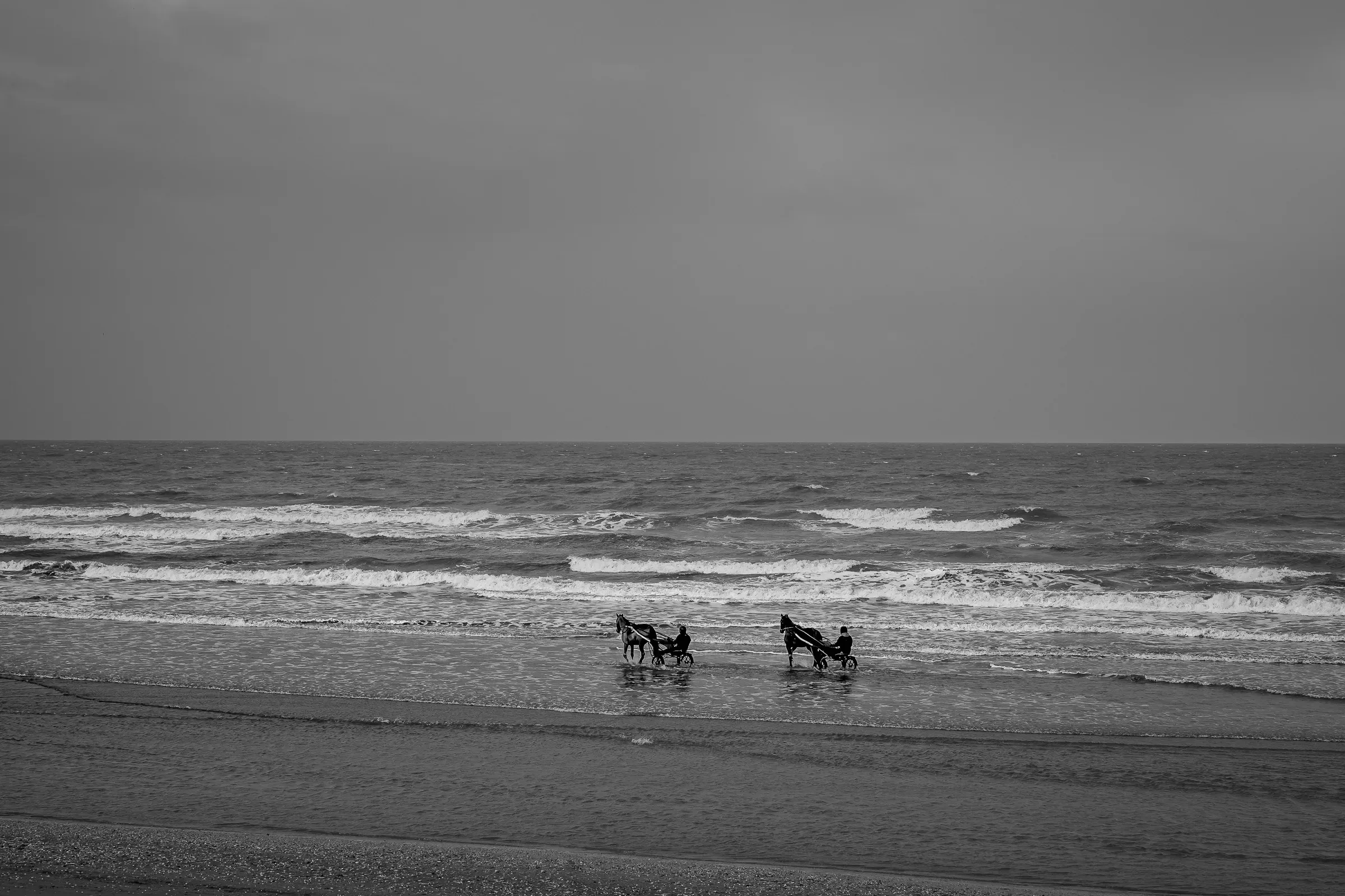 Two horse-drawn sulkies exercising on a flat beach, their silhouettes tiny against a wide grey sea and overcast sky