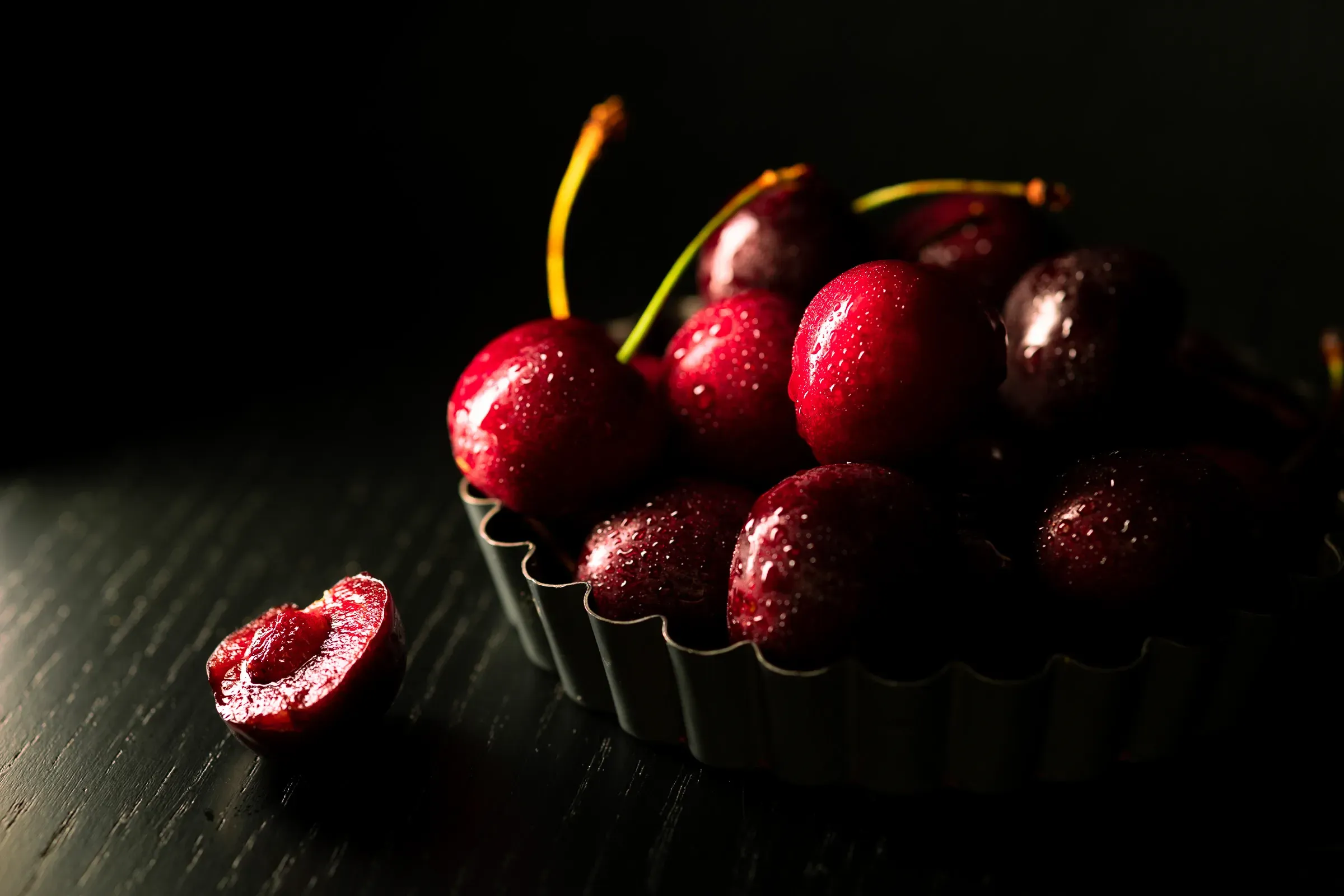 Dark cherries piled in a small metal fluted tart ring on a dark wooden surface, one cherry halved to show the pit, near-black background