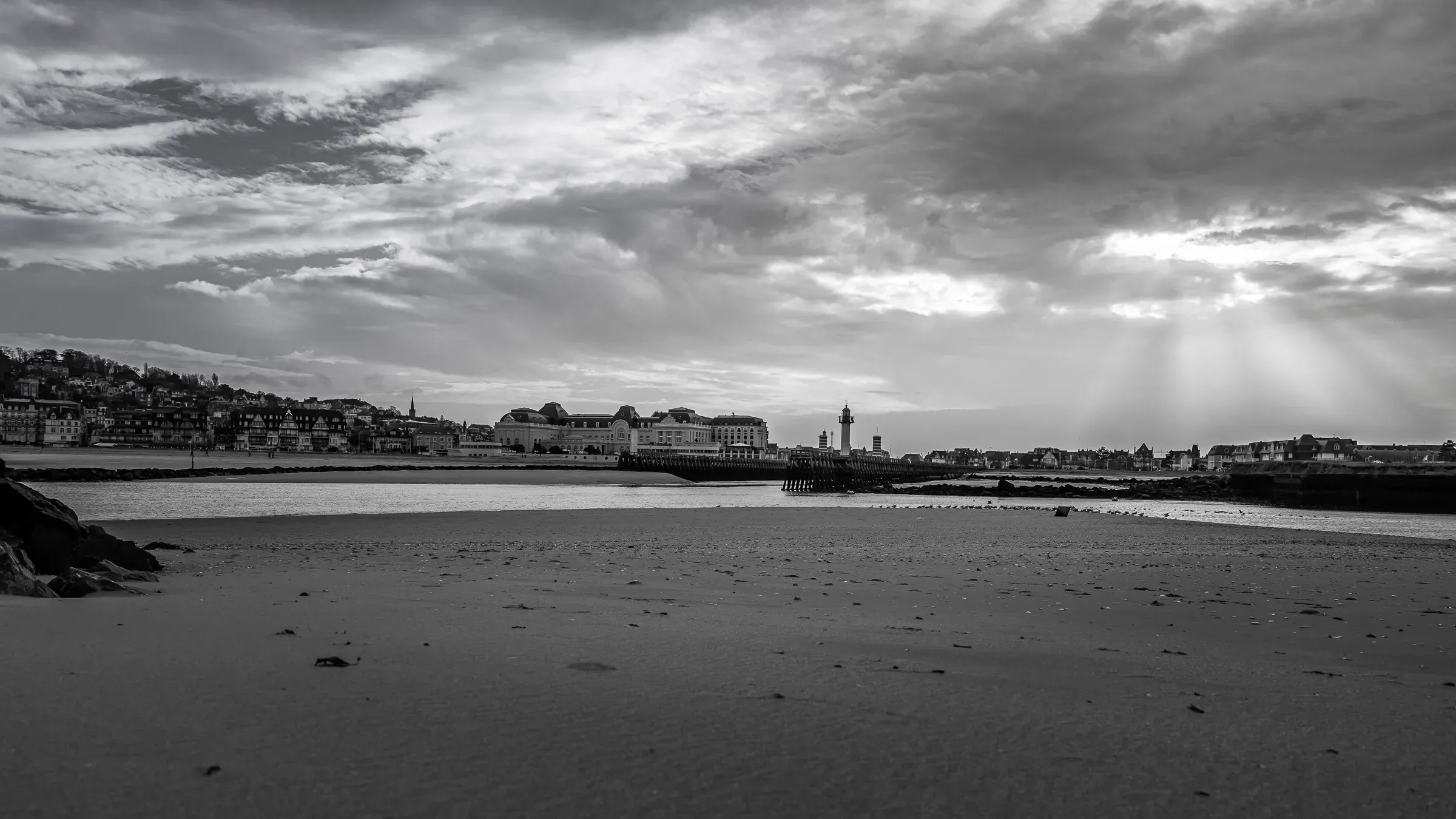 Low-tide beach at a Normandy coastal town, a lighthouse and seafront buildings on the horizon, sunrays breaking through clouds above