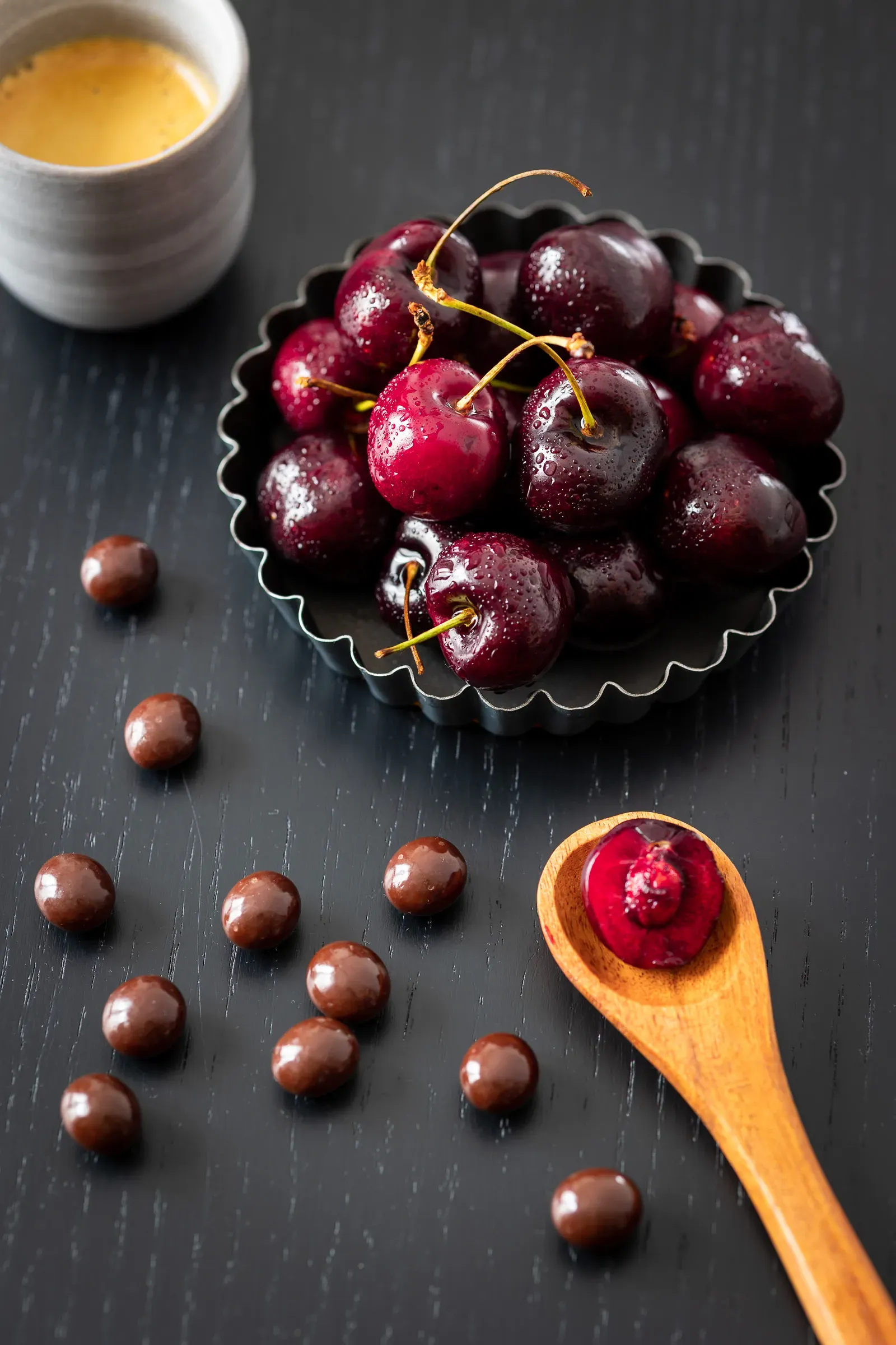 Dark cherries in a metal fluted ring, chocolate balls scattered on the dark surface beside it, a wooden spoon with a single cherry on top