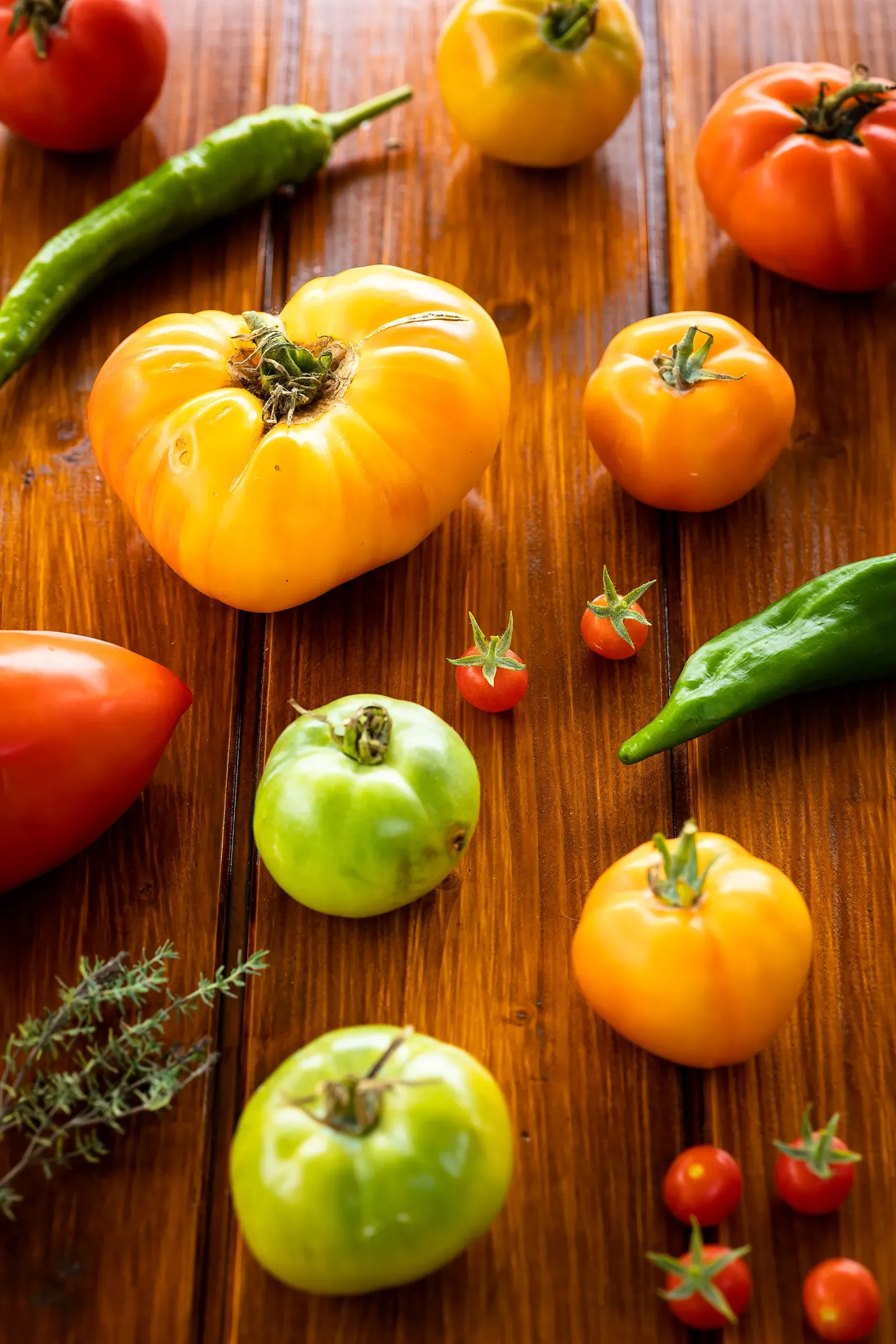 Heirloom tomatoes in red, orange, yellow and green scattered across a dark wood surface with green chili peppers and a sprig of thyme