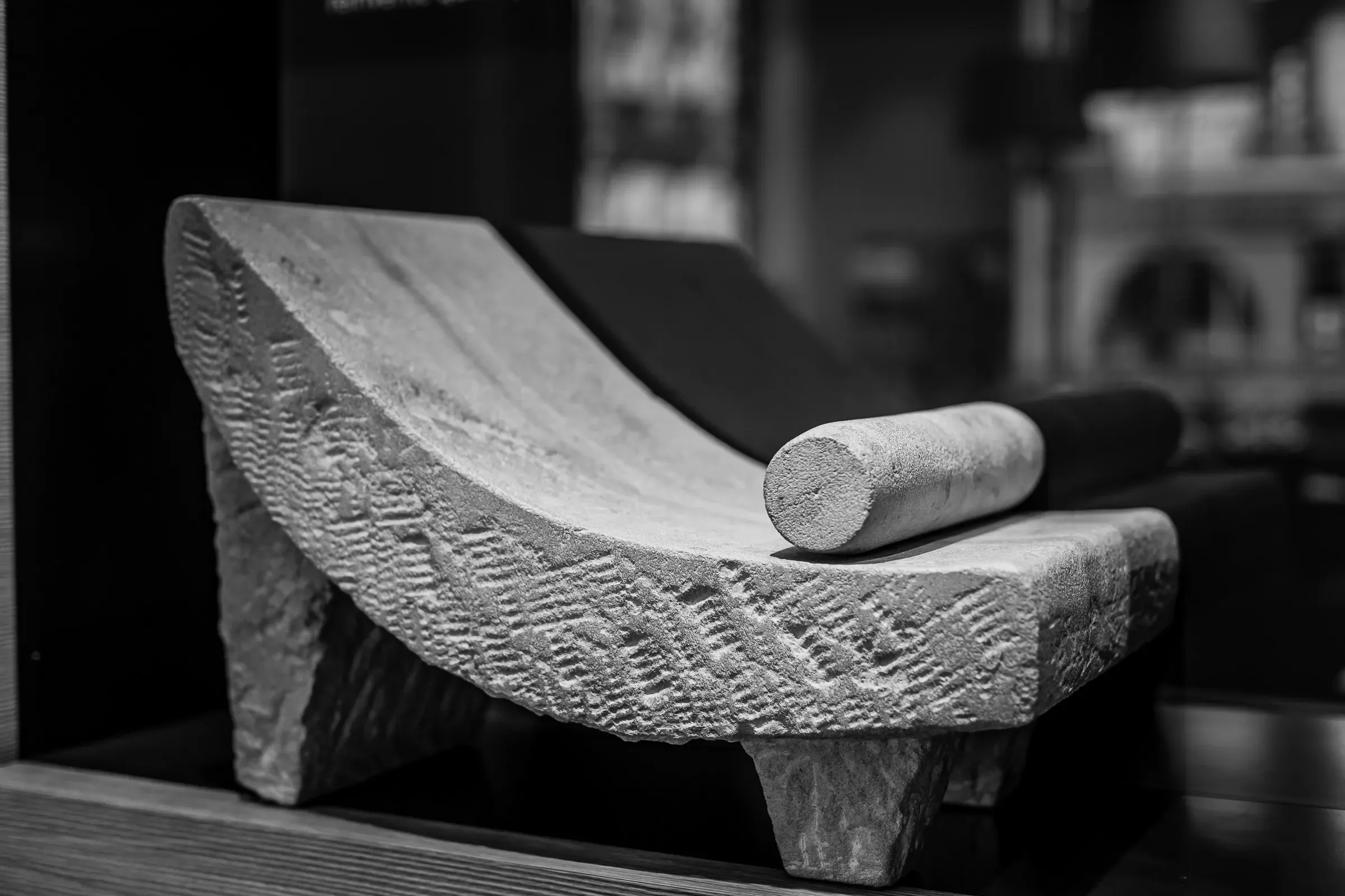Ancient stone headrest displayed in a museum vitrine, its curved surface incised with geometric chevron pattern, a cylindrical pillow beside it