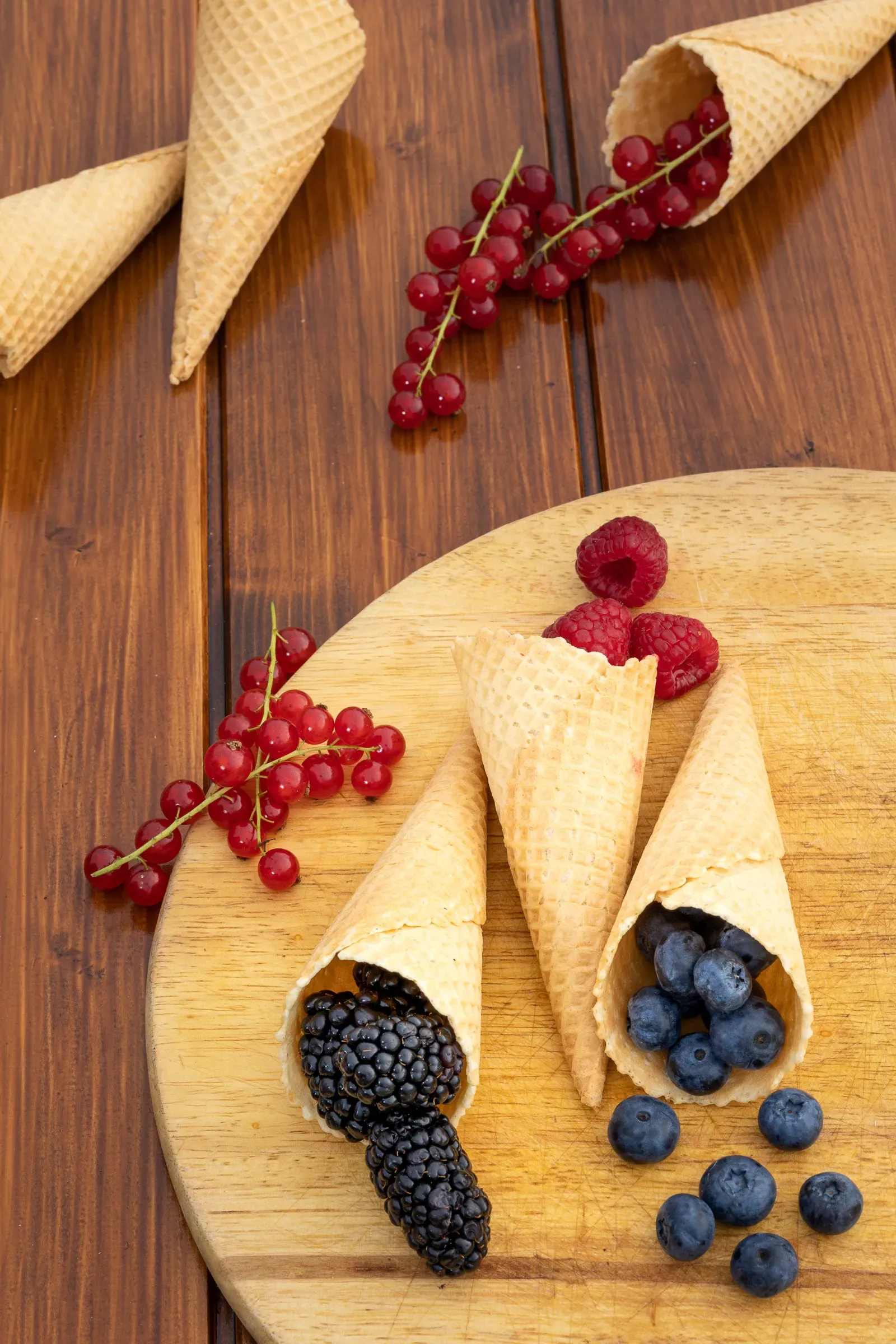 Waffle cones filled with blackberries, blueberries and raspberries on a round wooden board, red currant clusters beside them
