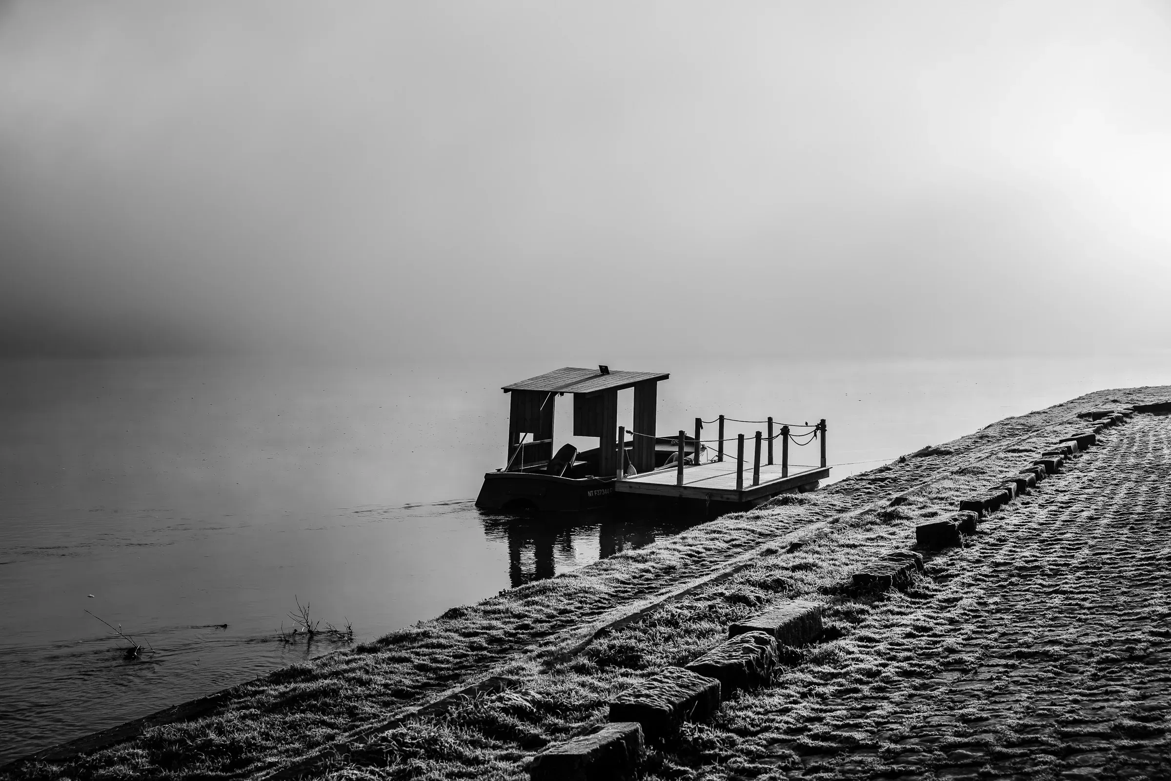 A small flat-bottomed ferry moored at a misty lakeside, frost covering the stone embankment, everything dissolving into grey fog