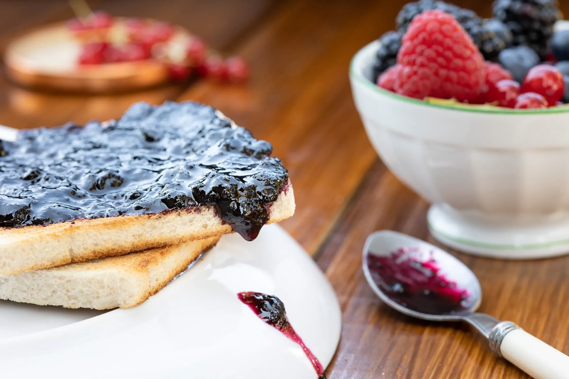 Toast thickly spread with dark mixed berry jam on a white plate, bowl of fresh berries and a spoon to the right on a wooden table