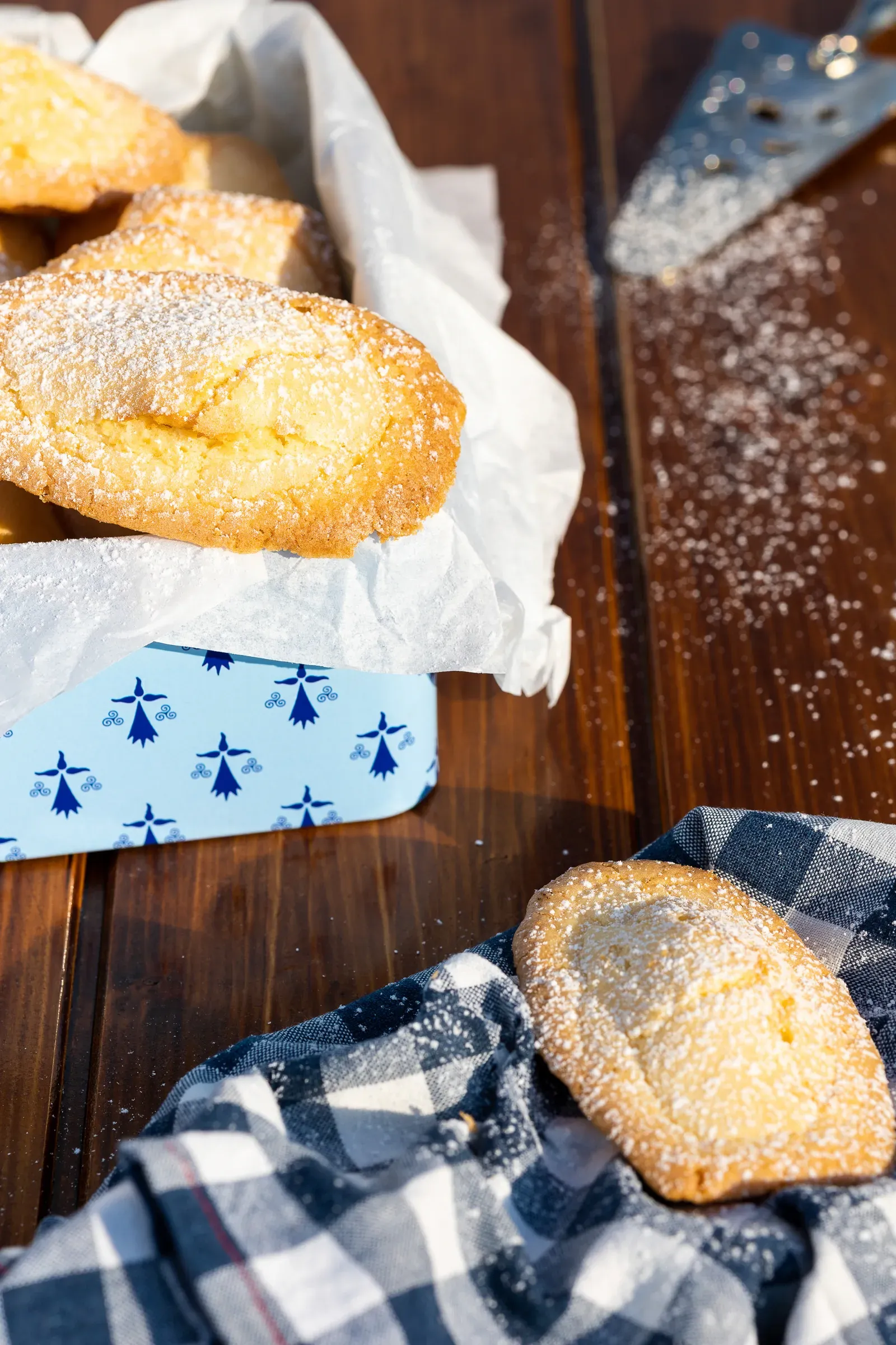 Madeleines dusted with powdered sugar in a blue-patterned decorative tin on a dark wood surface, one madeleine on a gingham cloth beside it