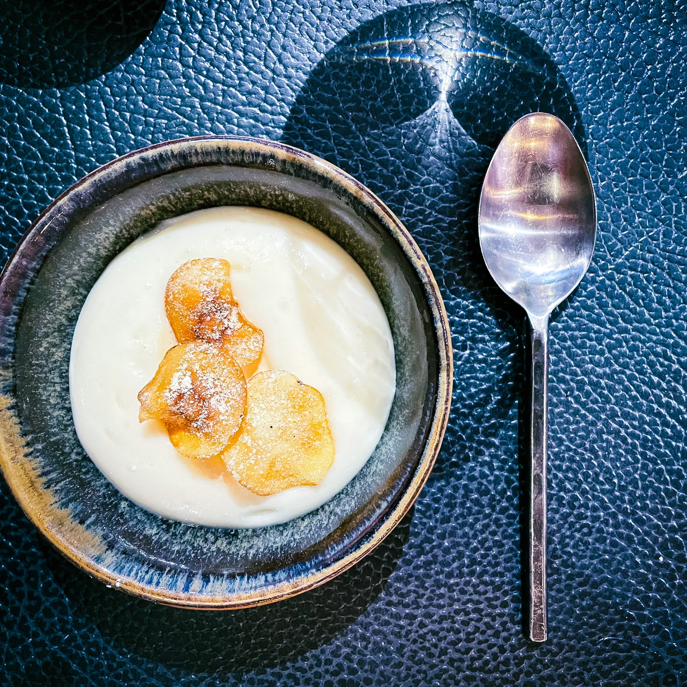 Rice pudding in a blue-grey ceramic bowl, two caramelised apple chips on top, silver spoon beside it on a dark textured surface