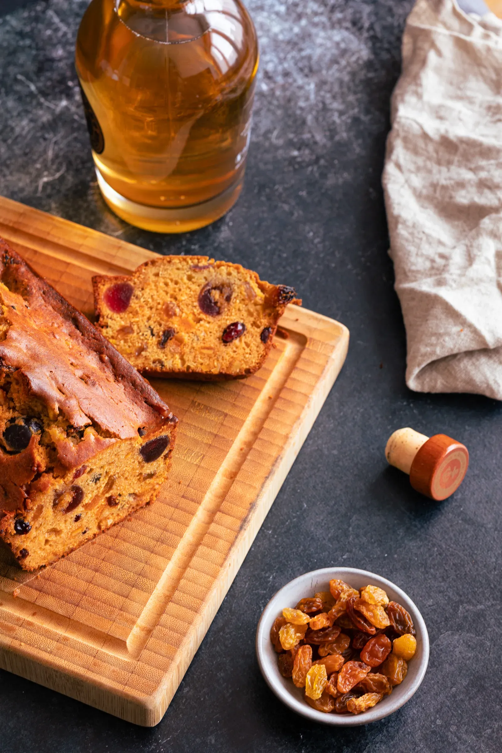 Rum fruit cake sliced on a wooden board — cherries and raisins visible in the dark crumb, a bottle of rum and bowl of golden raisins beside it