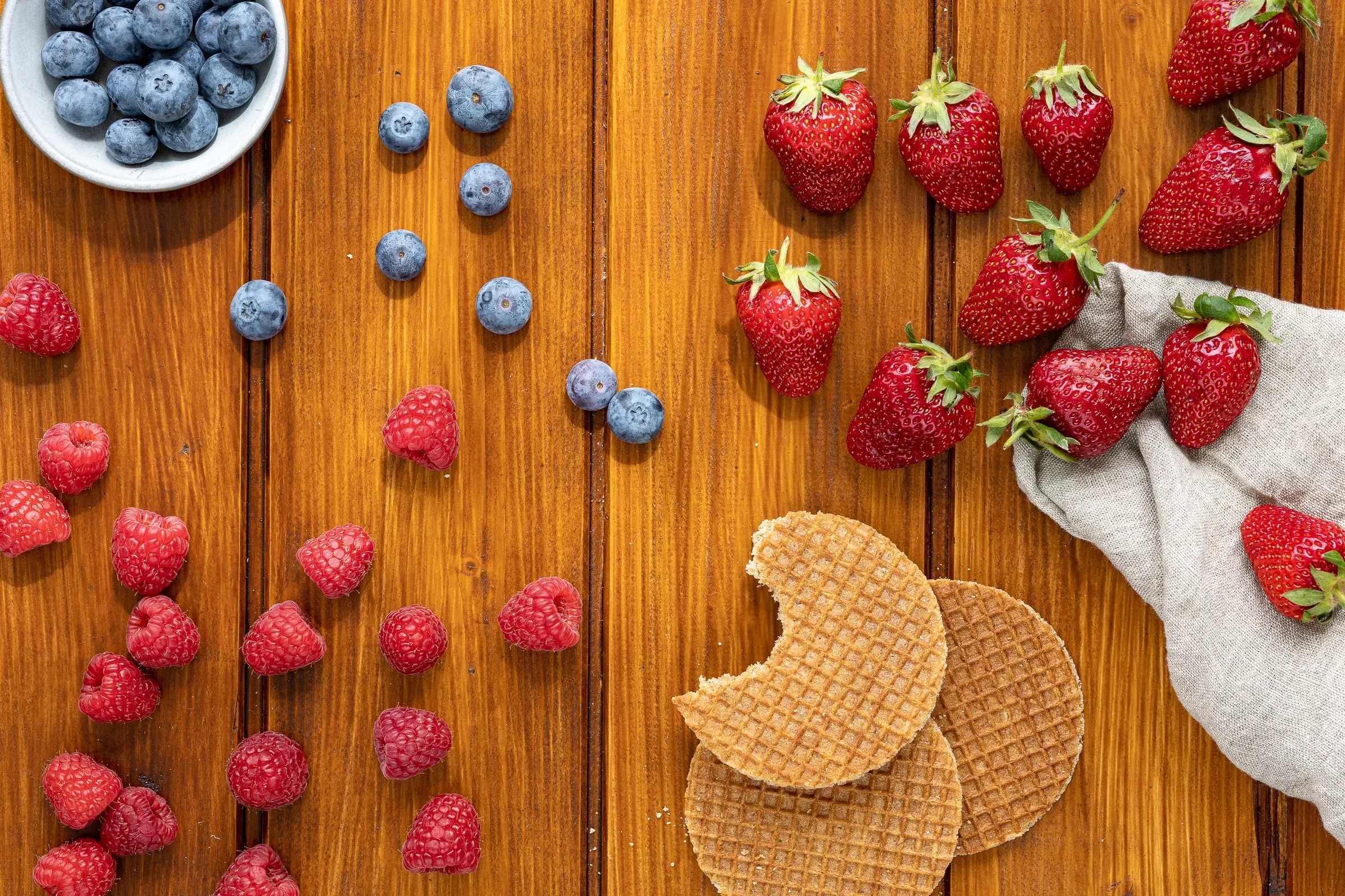 Overhead of blueberries, raspberries and strawberries scattered on dark wood beside two stroopwafels, a cloth napkin at the edge