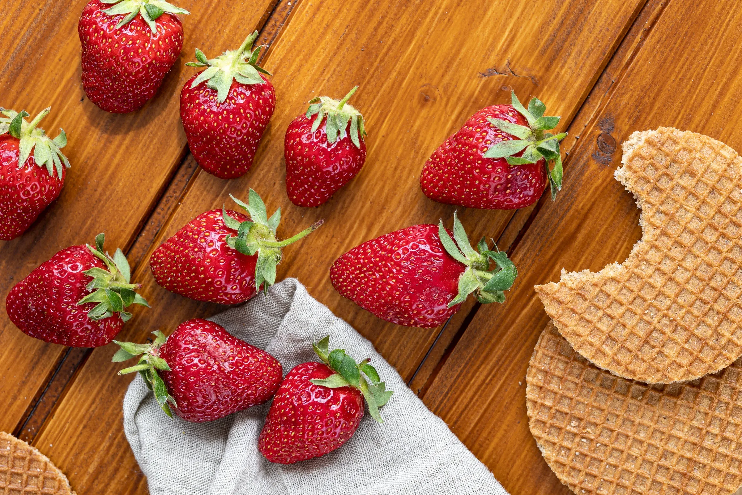 Strawberries spilling from a linen cloth beside broken stroopwafel pieces on a dark wood surface, closer crop