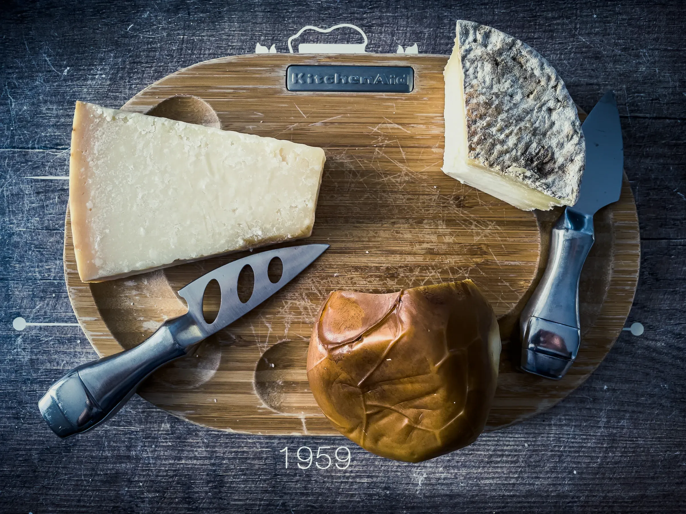 Cheese board overhead — a wedge of aged hard cheese, a round washed-rind, and a pressed cheese, two knives, dark wood surface