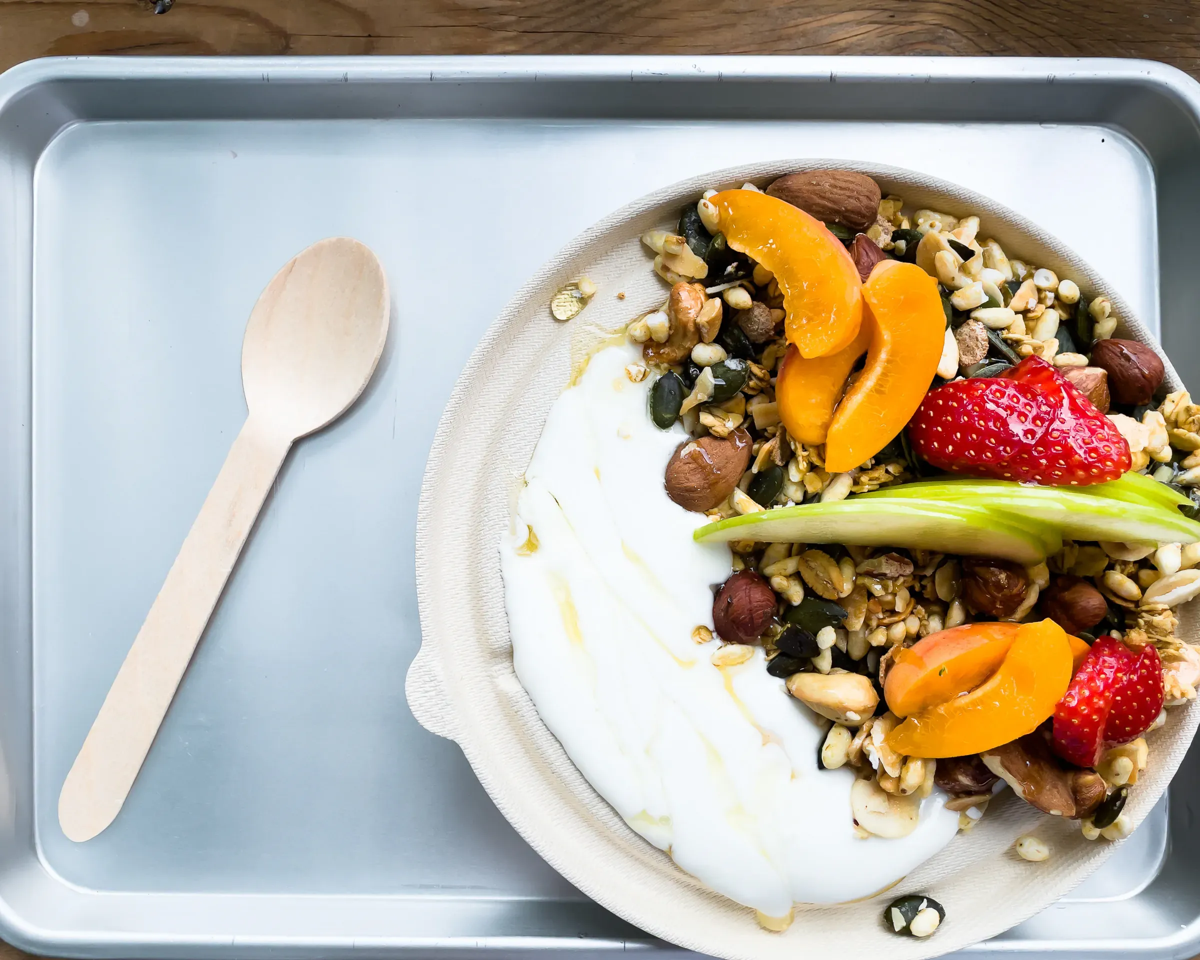 Granola bowl with yogurt, apricot slices, a strawberry, hazelnuts, honey drizzle and a green apple slice, wooden spoon on a metal tray