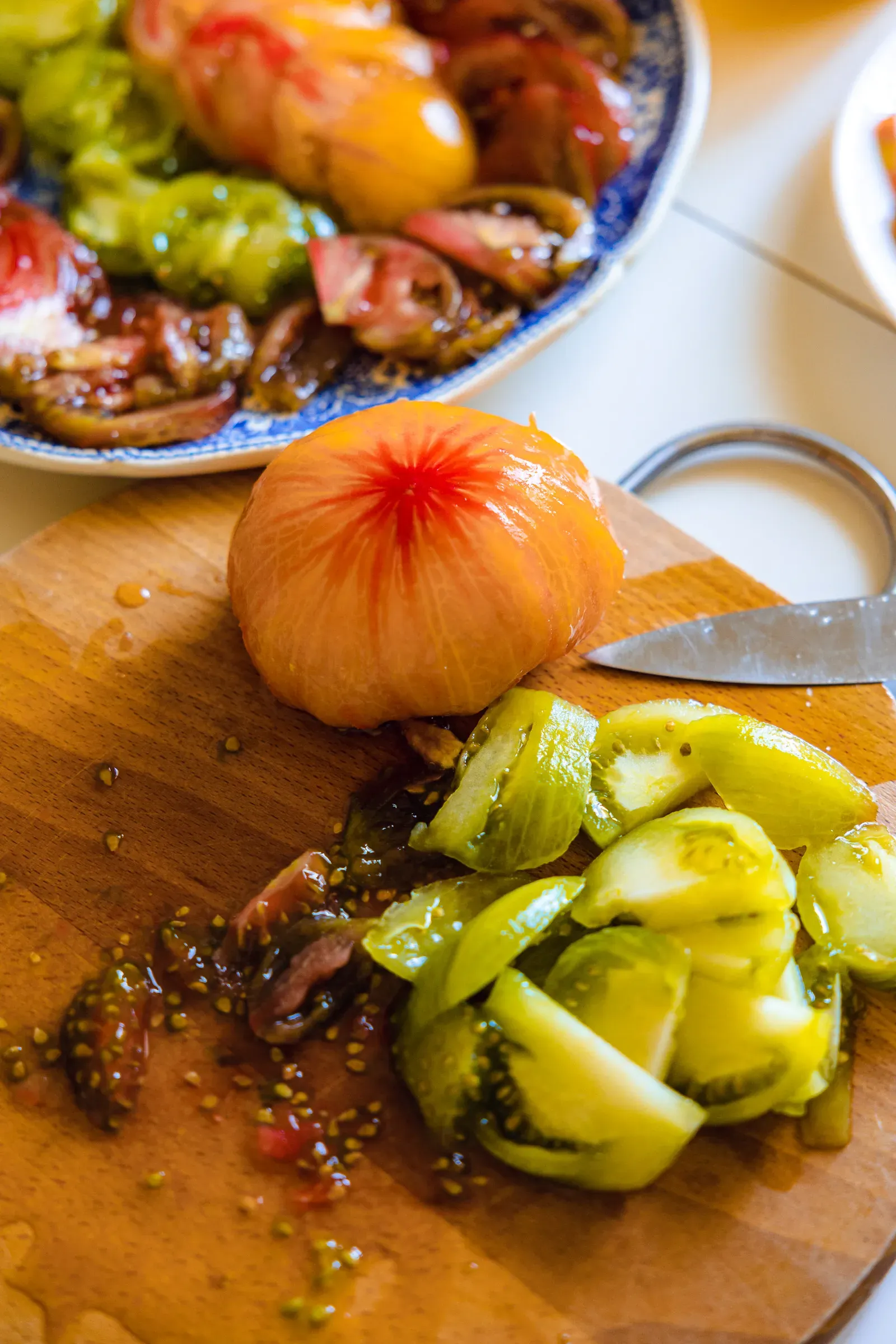 Heirloom tomato being sliced on a wooden board, green tomato wedges in the foreground, a bowl of tomato salad behind
