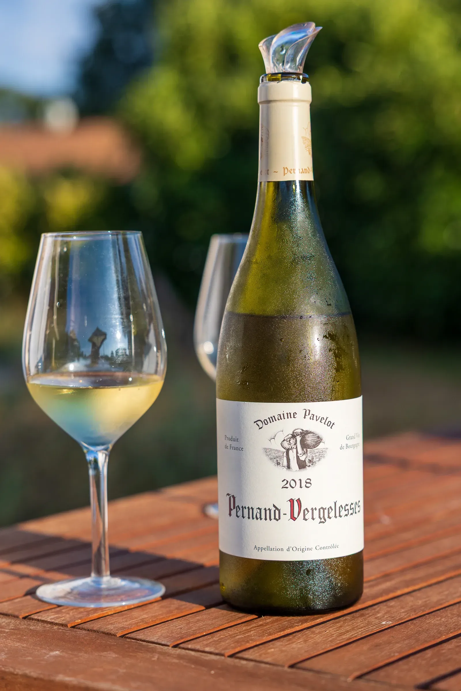 Bottle of Pernand-Vergelesses 2018, Domaine Pavelot, beside a glass of white wine on a wooden garden table, soft green bokeh behind