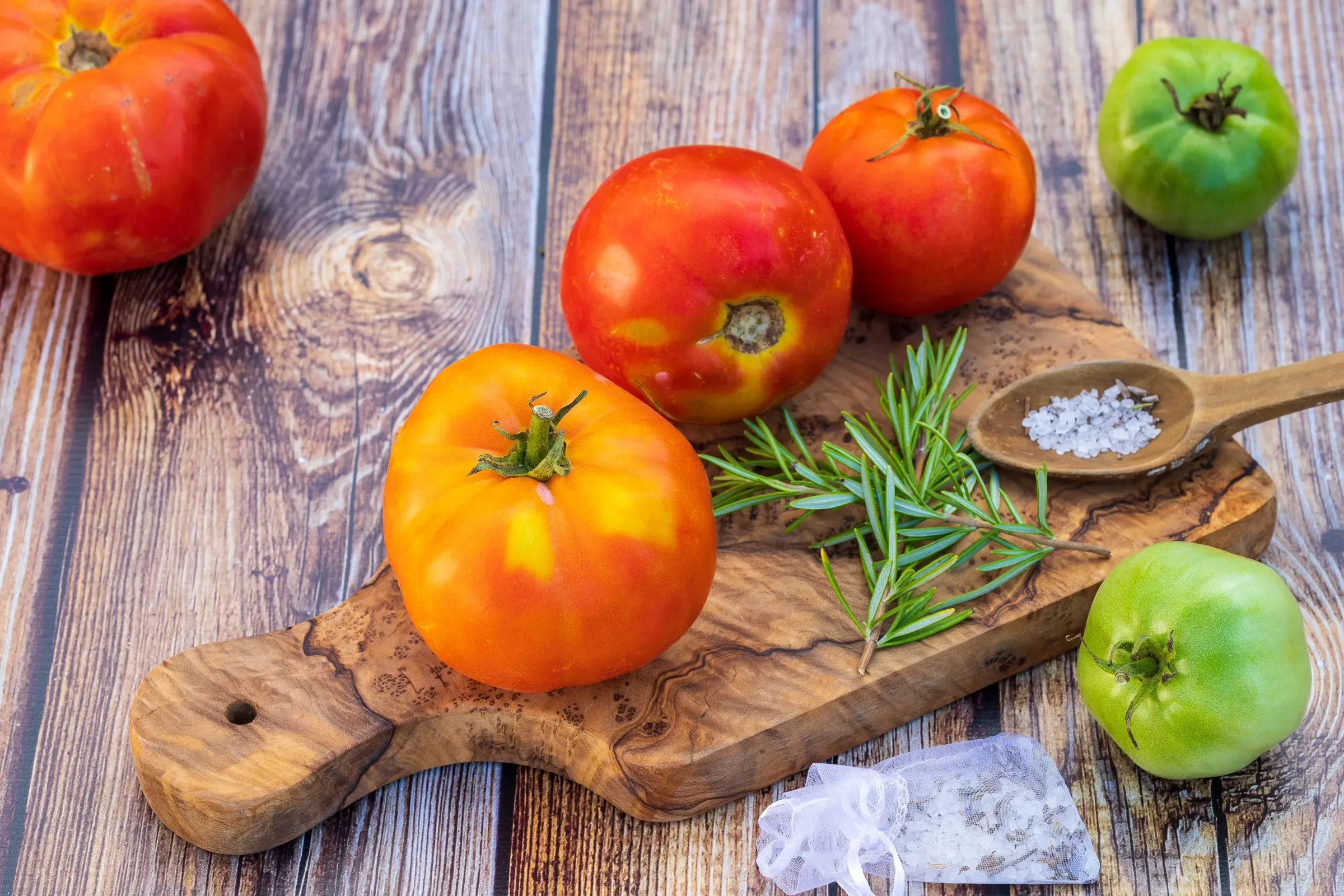 Heirloom tomatoes on an olive wood board with a sprig of rosemary and a spoon of sea salt, two green tomatoes on the dark wood surface