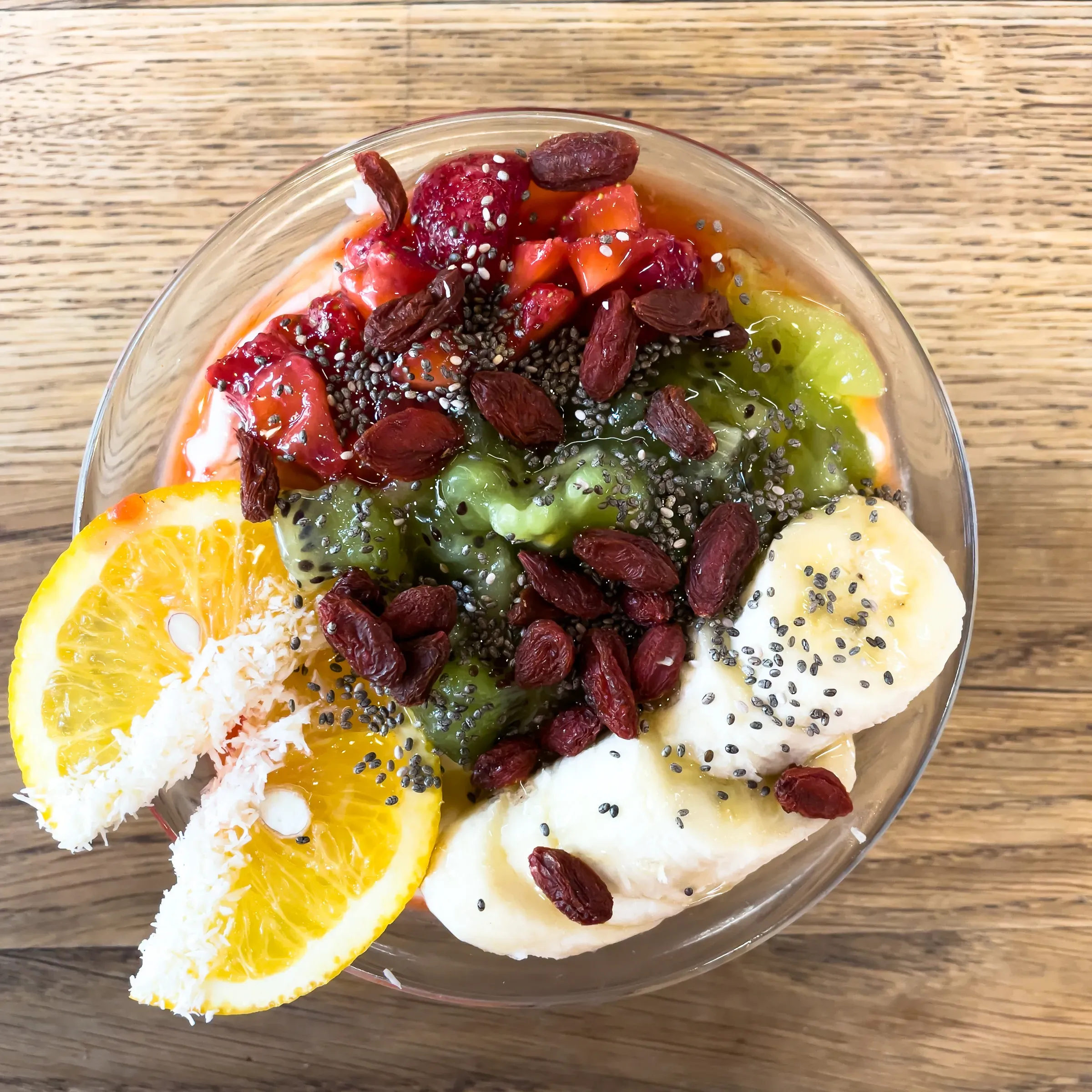 Acai smoothie bowl overhead — kiwi, banana slices, orange round, strawberries, goji berries, chia seeds and coconut on a wooden surface