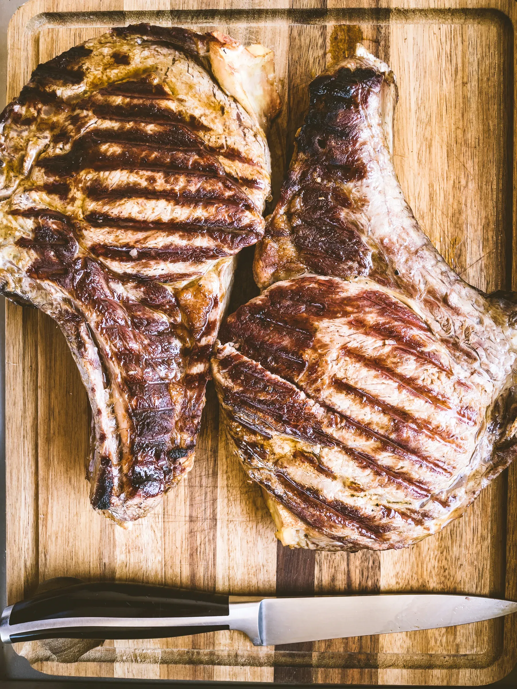 Two grilled bone-in ribeye steaks overhead on a wooden cutting board, char marks visible, carving knife resting below