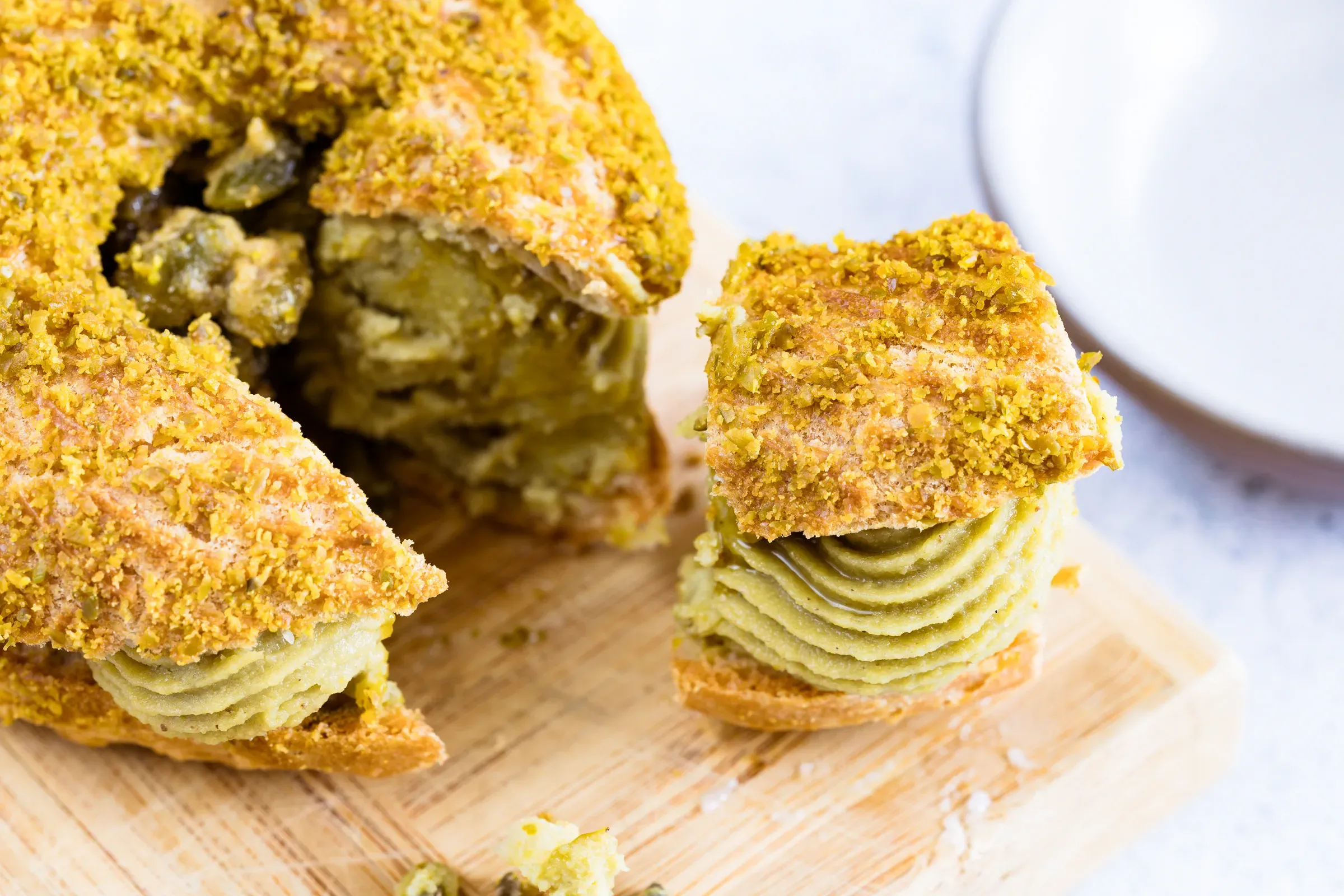 Pistachio Paris-Brest cut open on a wooden board, green pistachio mousseline cream filling exposed, crumble topping on the crust