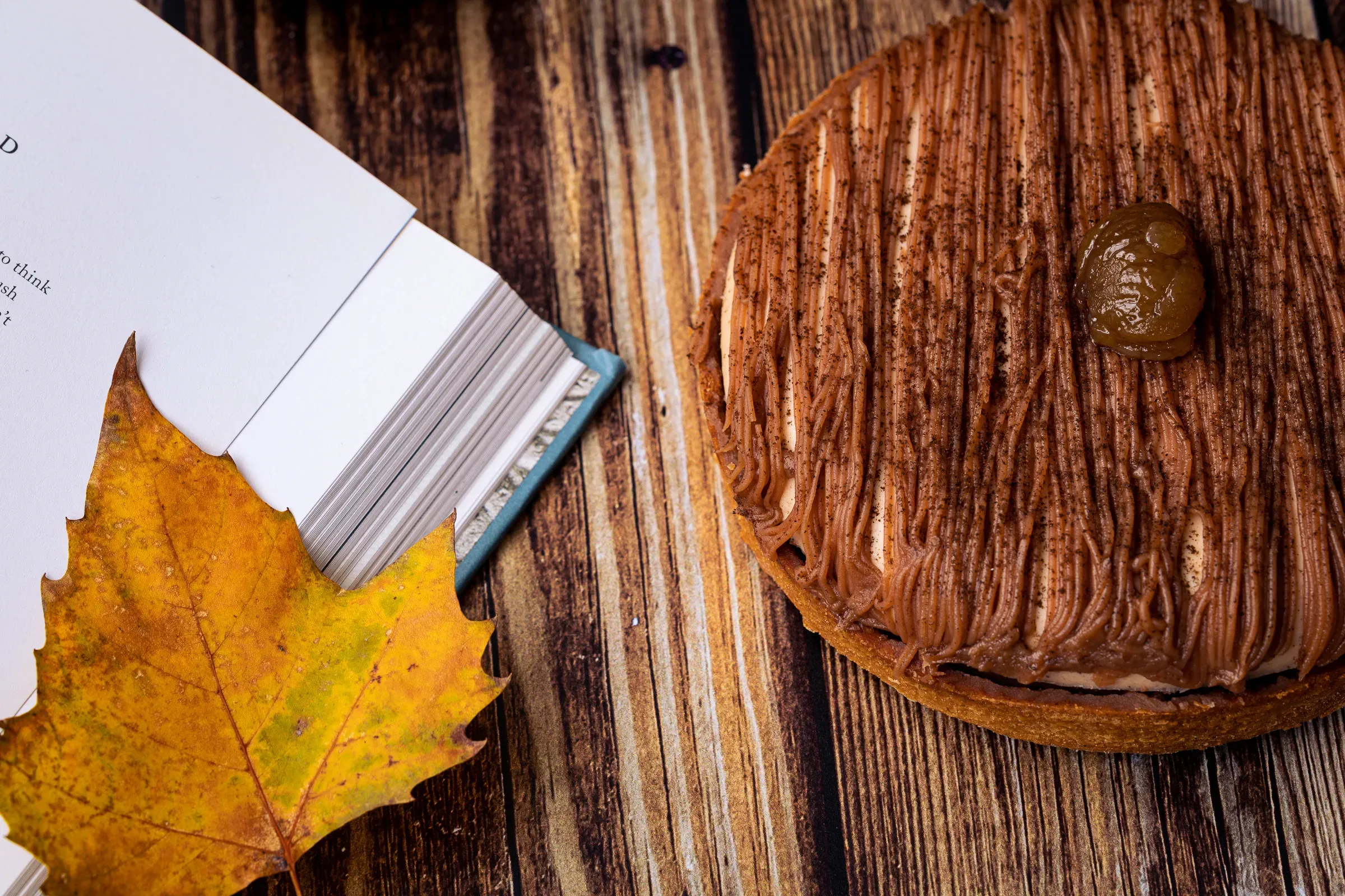 Mont Blanc tart close-up on a wooden surface, chestnut cream piped in fine strands, a yellow autumn leaf and open book beside it