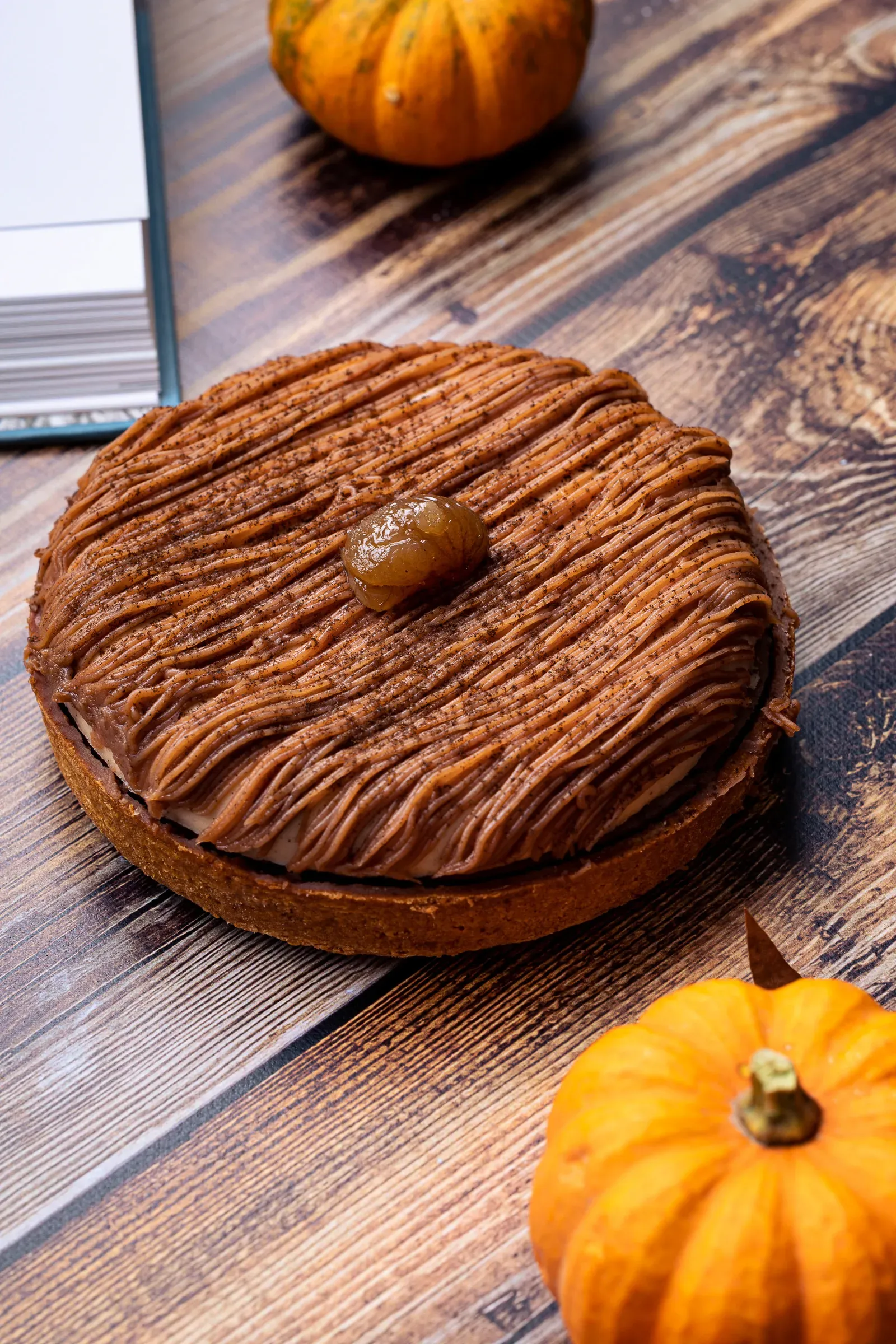 Mont Blanc tart on a wooden surface flanked by two small pumpkins, a book in the top left corner, vertical composition