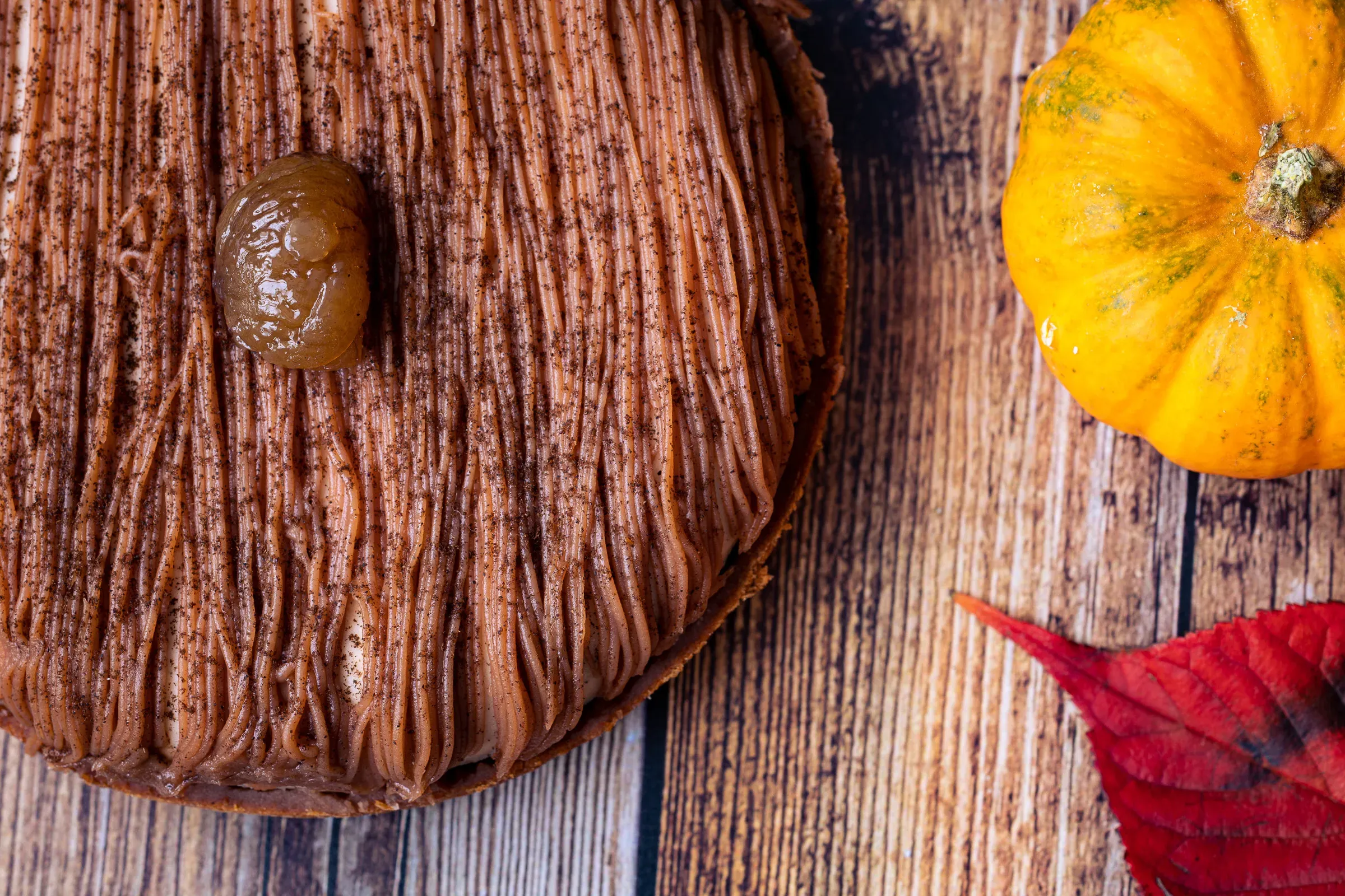 Mont Blanc tart overhead close-up — chestnut cream strands and a glazed whole chestnut at the centre, small pumpkin and red leaf at the edge