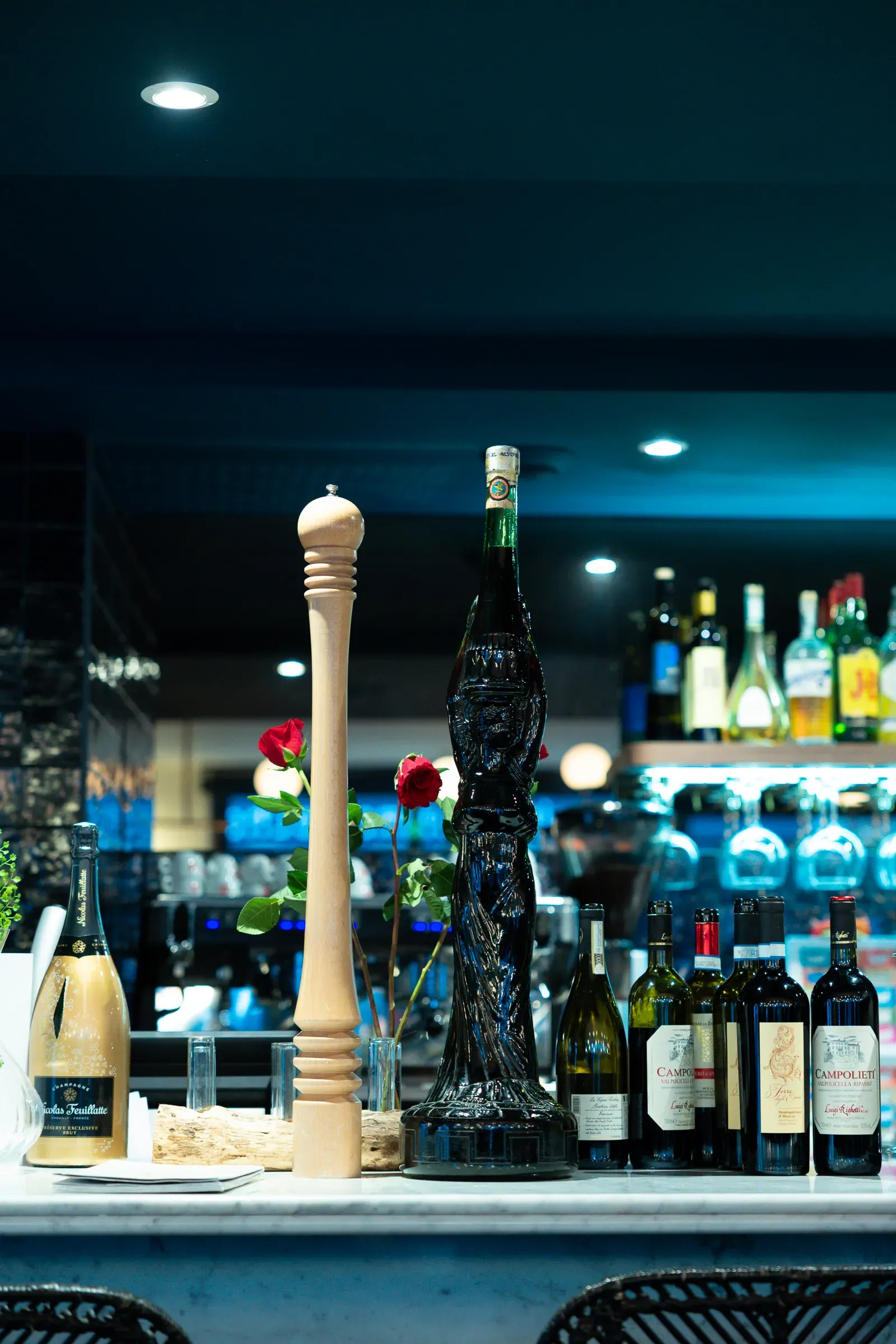 Restaurant bar counter — a tall pepper grinder, a dark sculptural bottle, wine bottles and red roses, backlit bottles on shelves behind