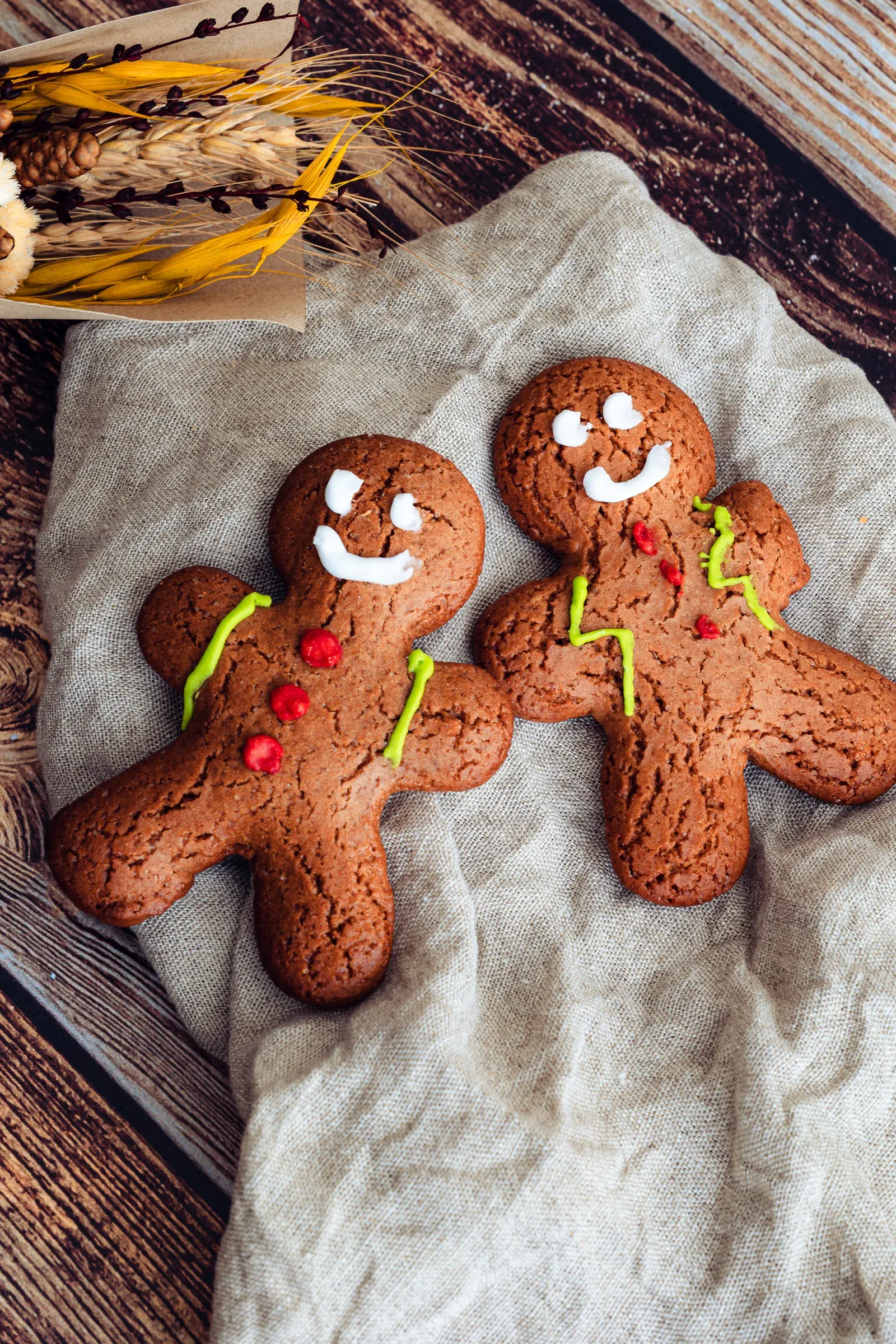 Two gingerbread men side by side on a linen cloth, white and green icing details, dried wheat stalks behind on a dark wooden surface