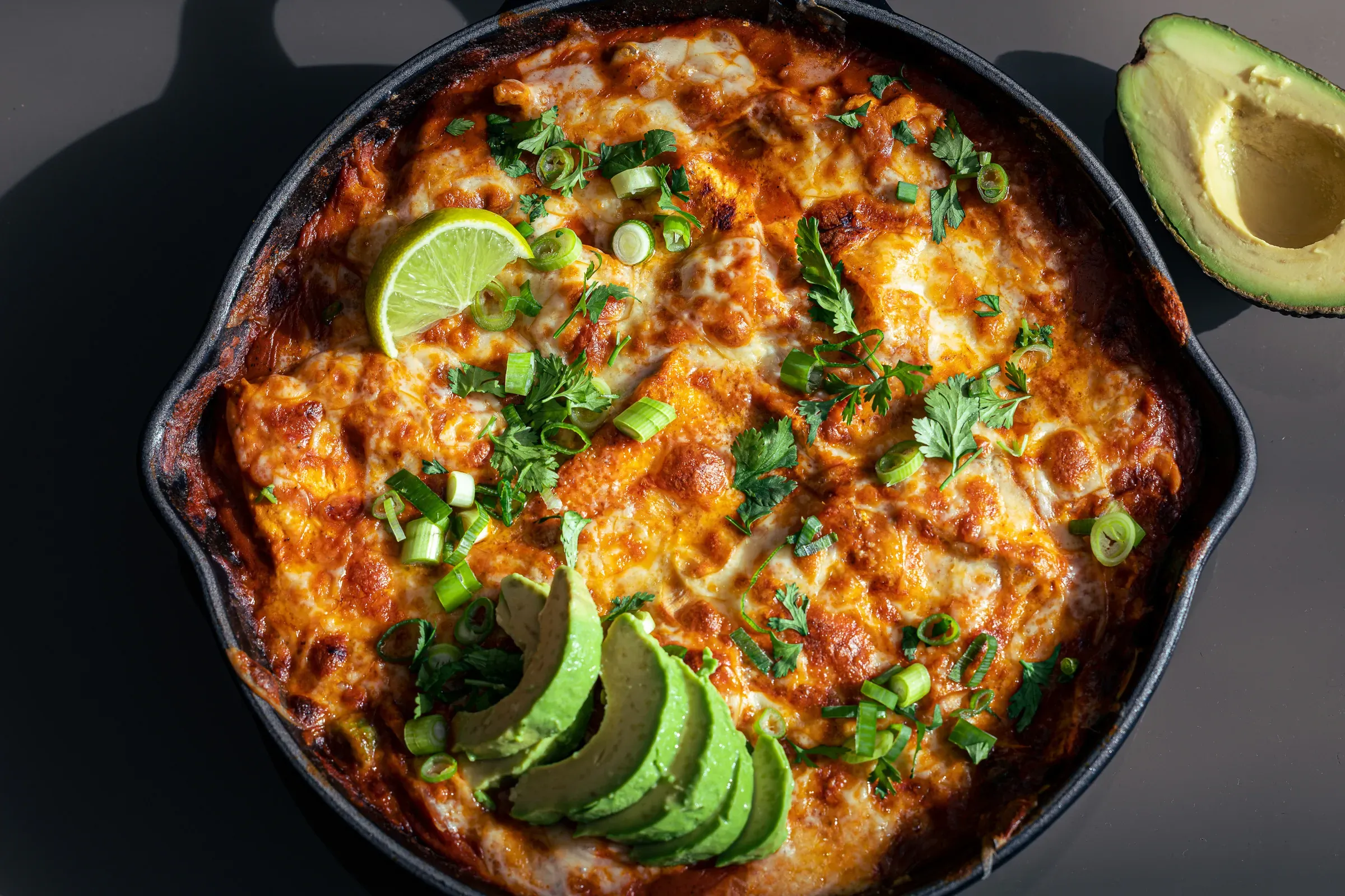 Enchiladas in a cast iron skillet — melted cheese browned at the edges, cilantro, sliced green onion, a lime wheel, half avocado beside