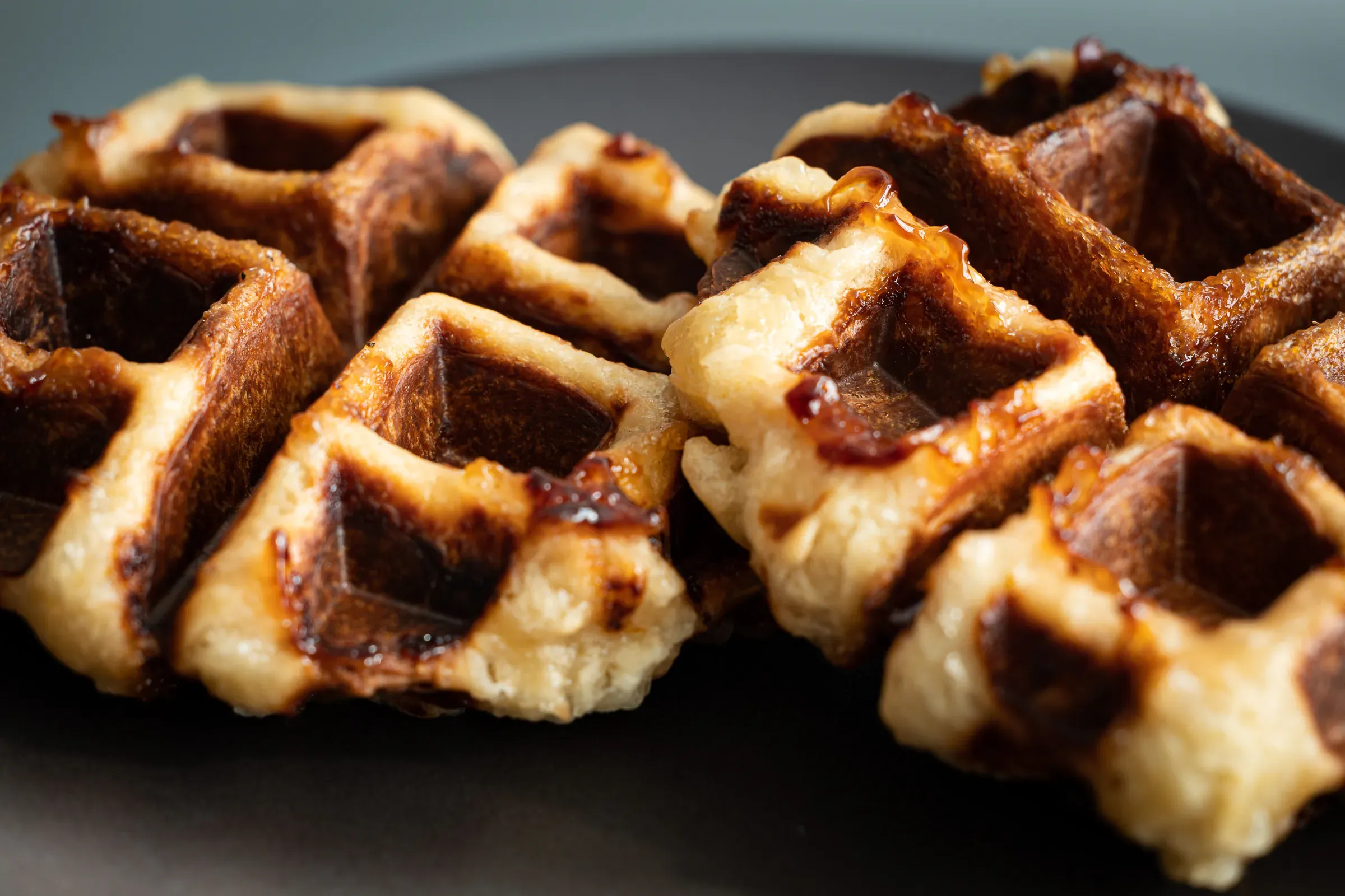 Two Liège waffles close-up on a dark plate, caramelised pearl sugar pockets dark and sticky at the surface