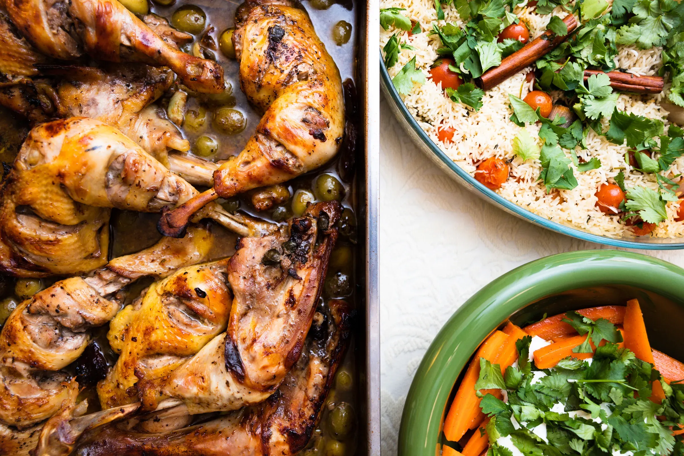Roasted chicken with green olives on a baking tray beside a bowl of spiced couscous with herbs and a carrot and cilantro salad