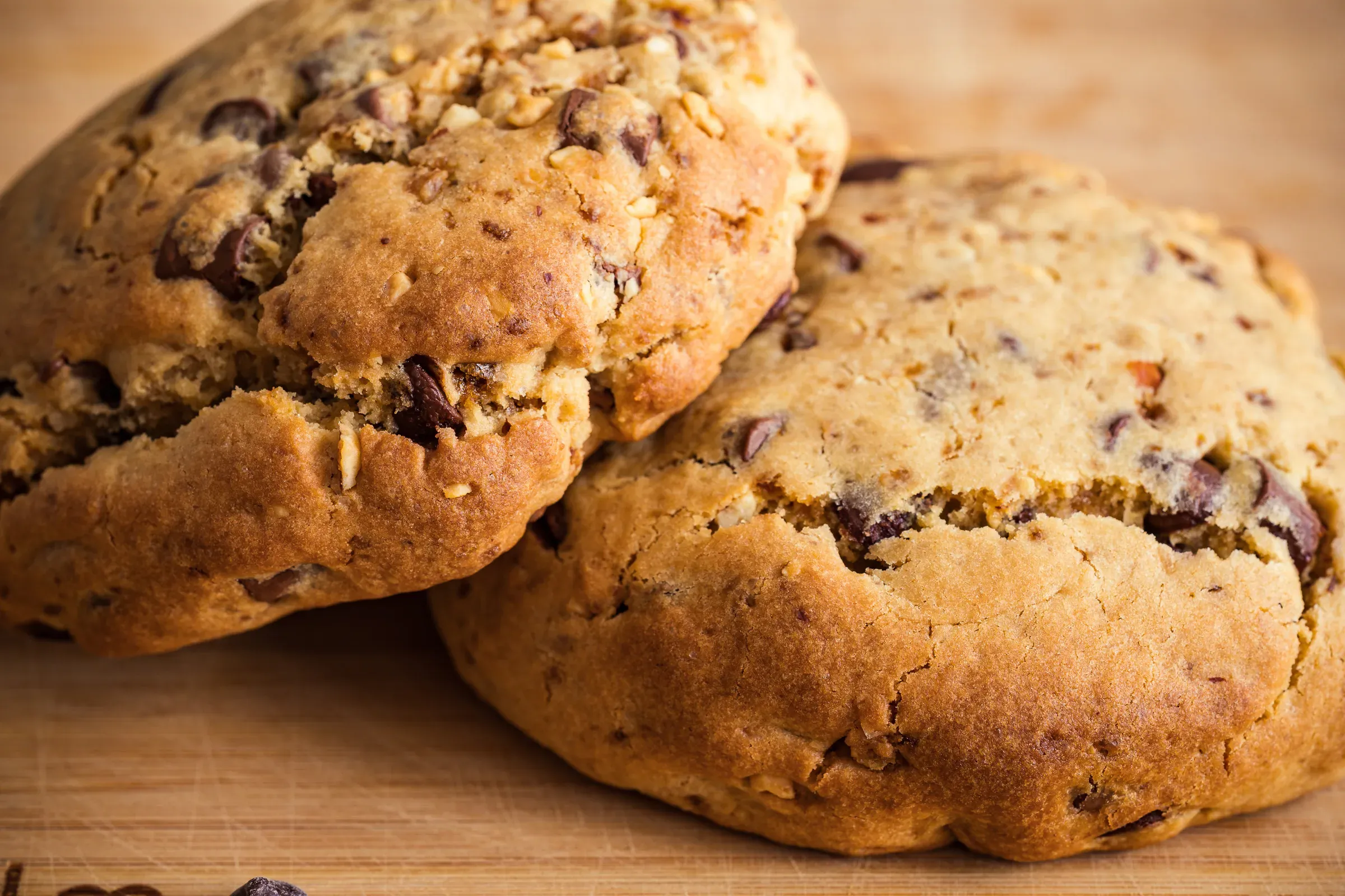 Three large chocolate chip cookies close-up on a wooden board, rough cracked texture, chocolate chunks visible