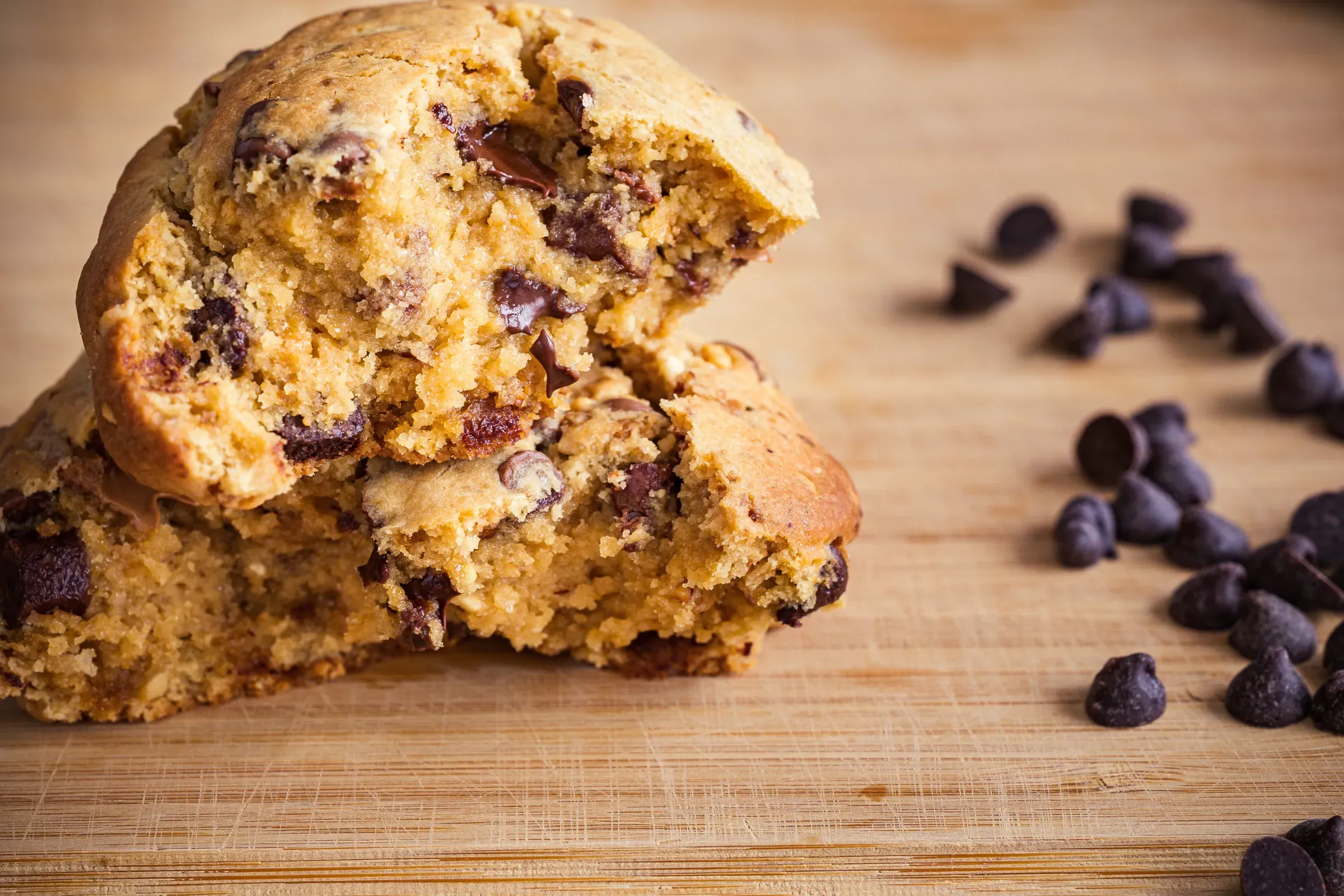 Chocolate chip cookie broken in two on a wooden board, soft interior and melted chocolate chunks visible, chips scattered beside it