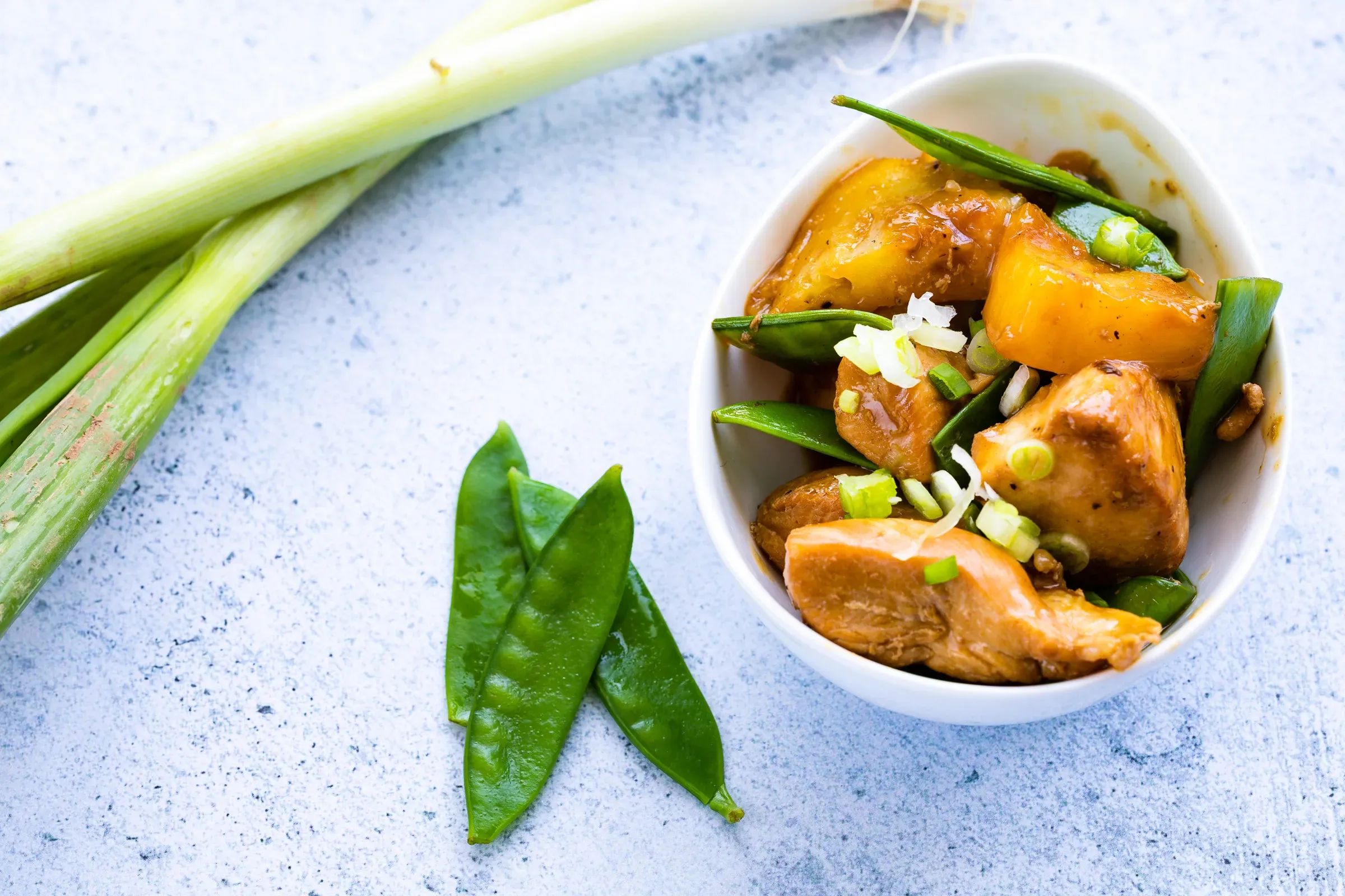 Sweet and sour chicken with mango chunks and snap peas in a white oval bowl, sliced spring onion and sesame on top, light blue surface
