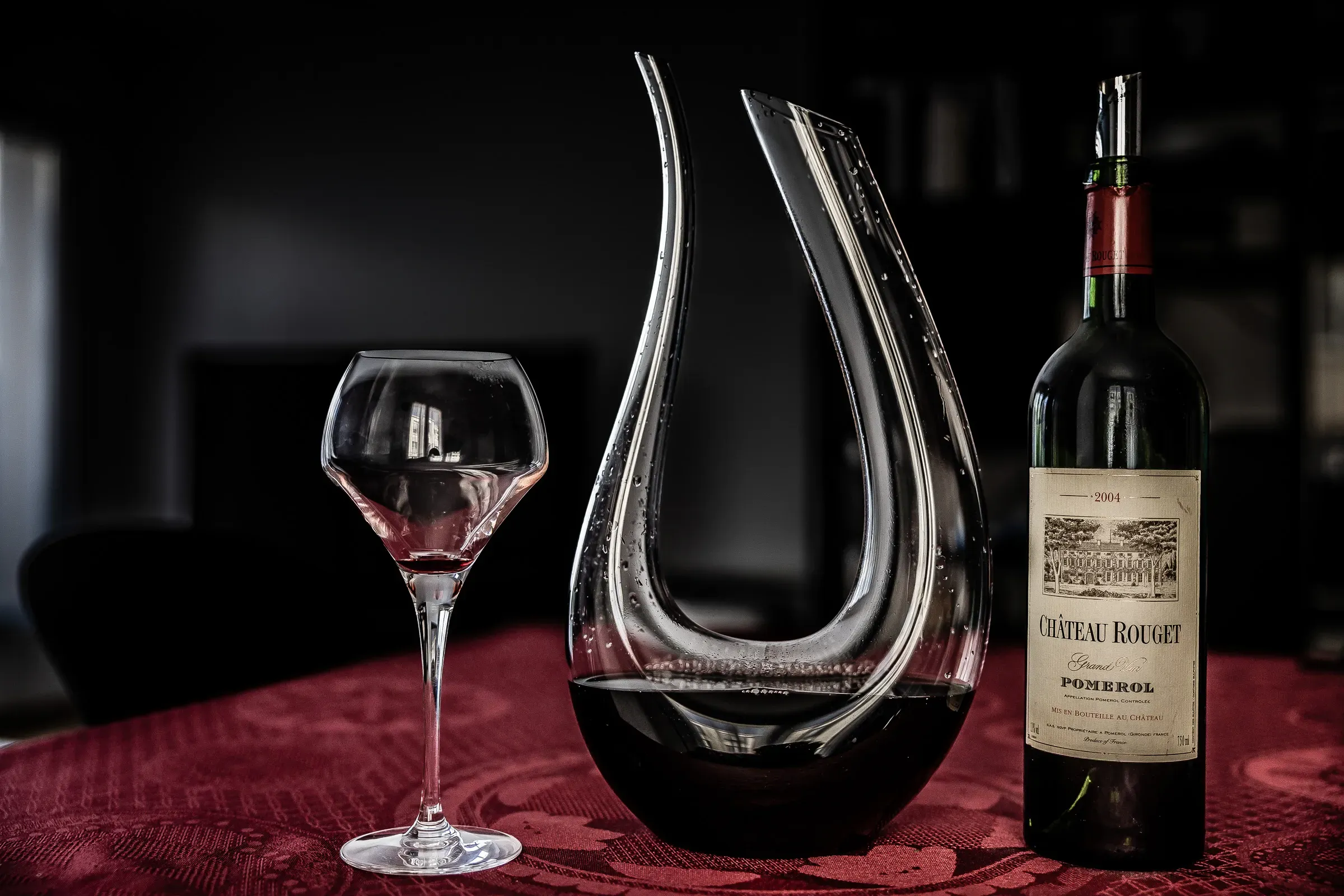 Château Rouget Pomerol bottle beside a swan-neck crystal decanter and a stemmed wine glass on a dark draped table, black background