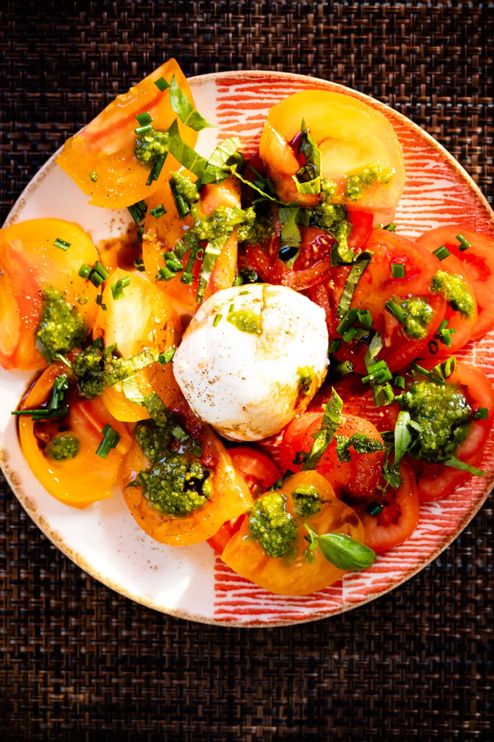 Heirloom tomato and burrata salad overhead on a patterned plate — orange and red tomato slices, a burrata ball, green pesto and chopped chives