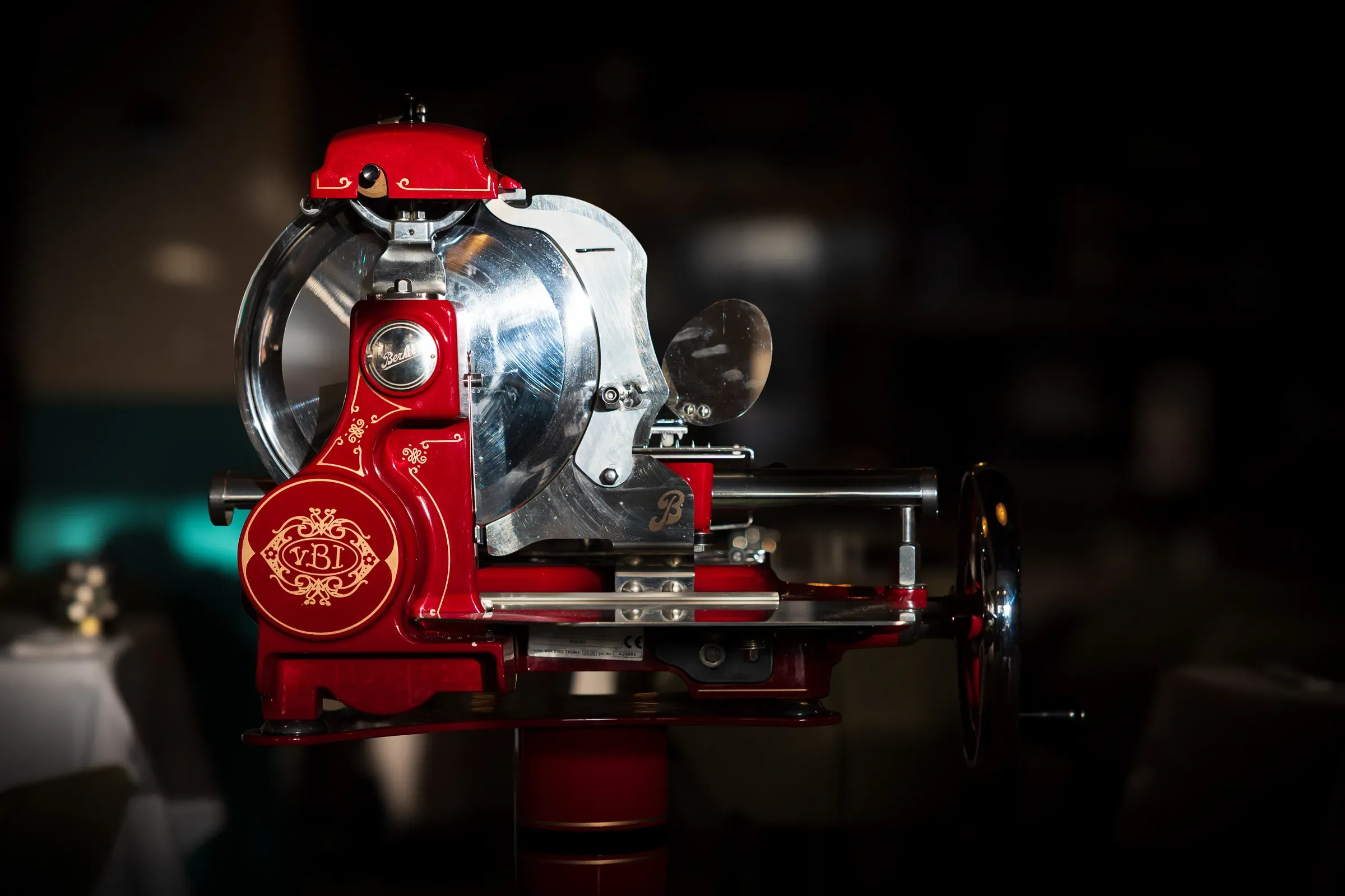 Vintage red Berkel hand-cranked meat slicer in profile, chrome blade and flywheel, gold script on the body, dark restaurant interior behind