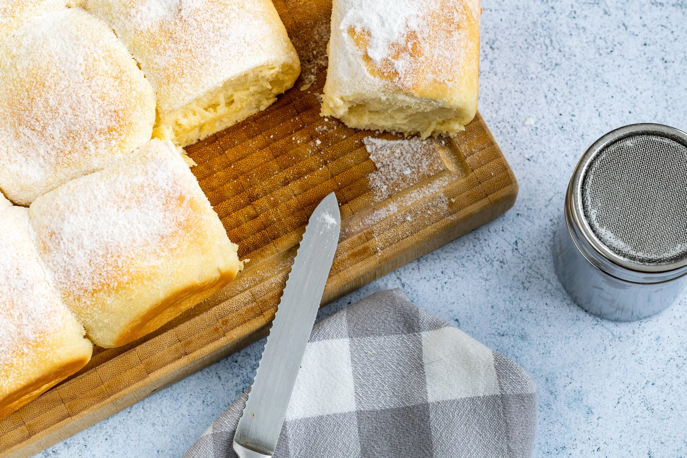 Pillowy milk bread rolls dusted with powdered sugar on a wooden board, a knife and grey checked cloth beside them, a sifter to the right