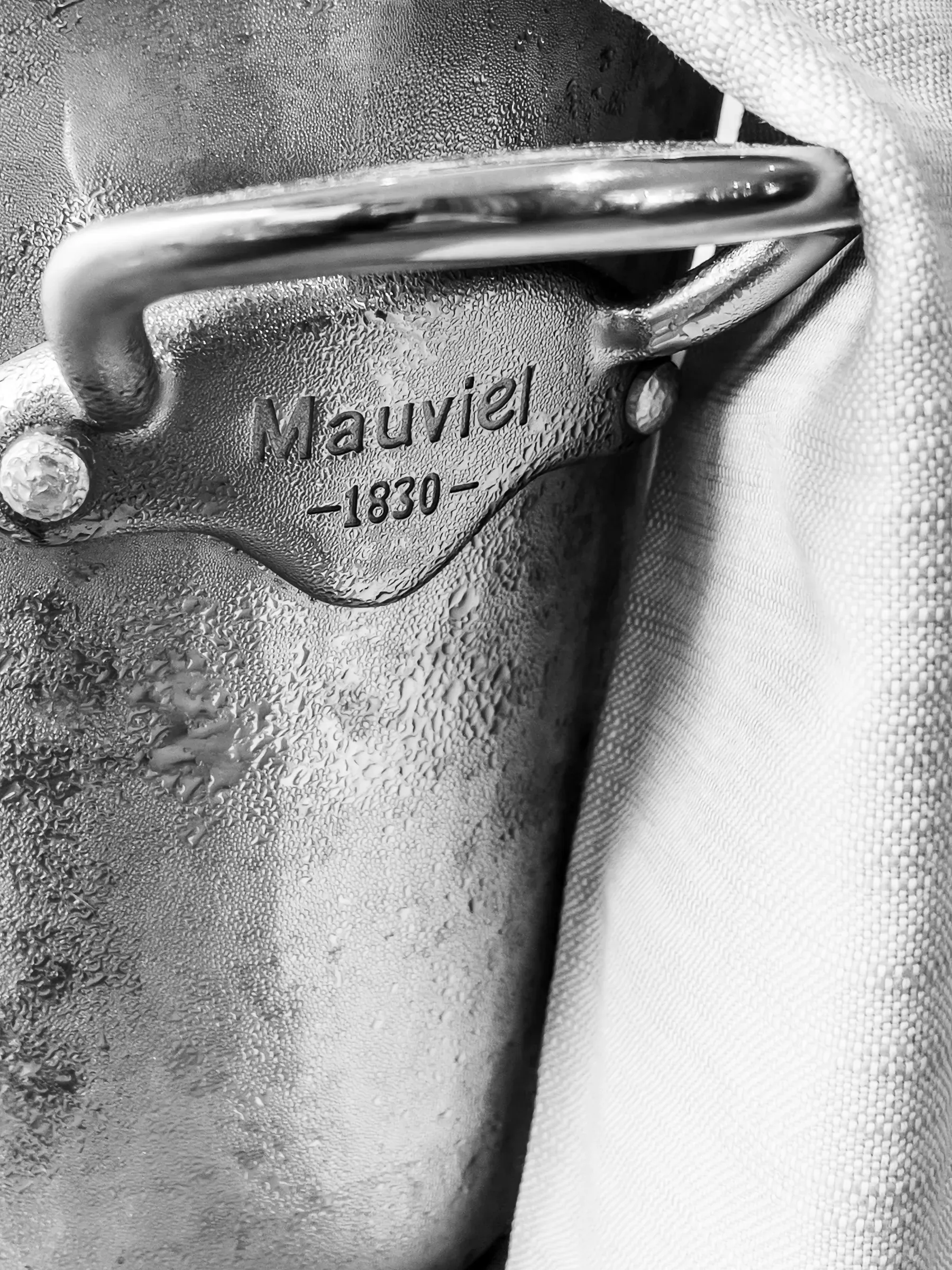 Mauviel 1830 copper pan handle close-up in black and white, condensation beading on the metal, white linen cloth behind