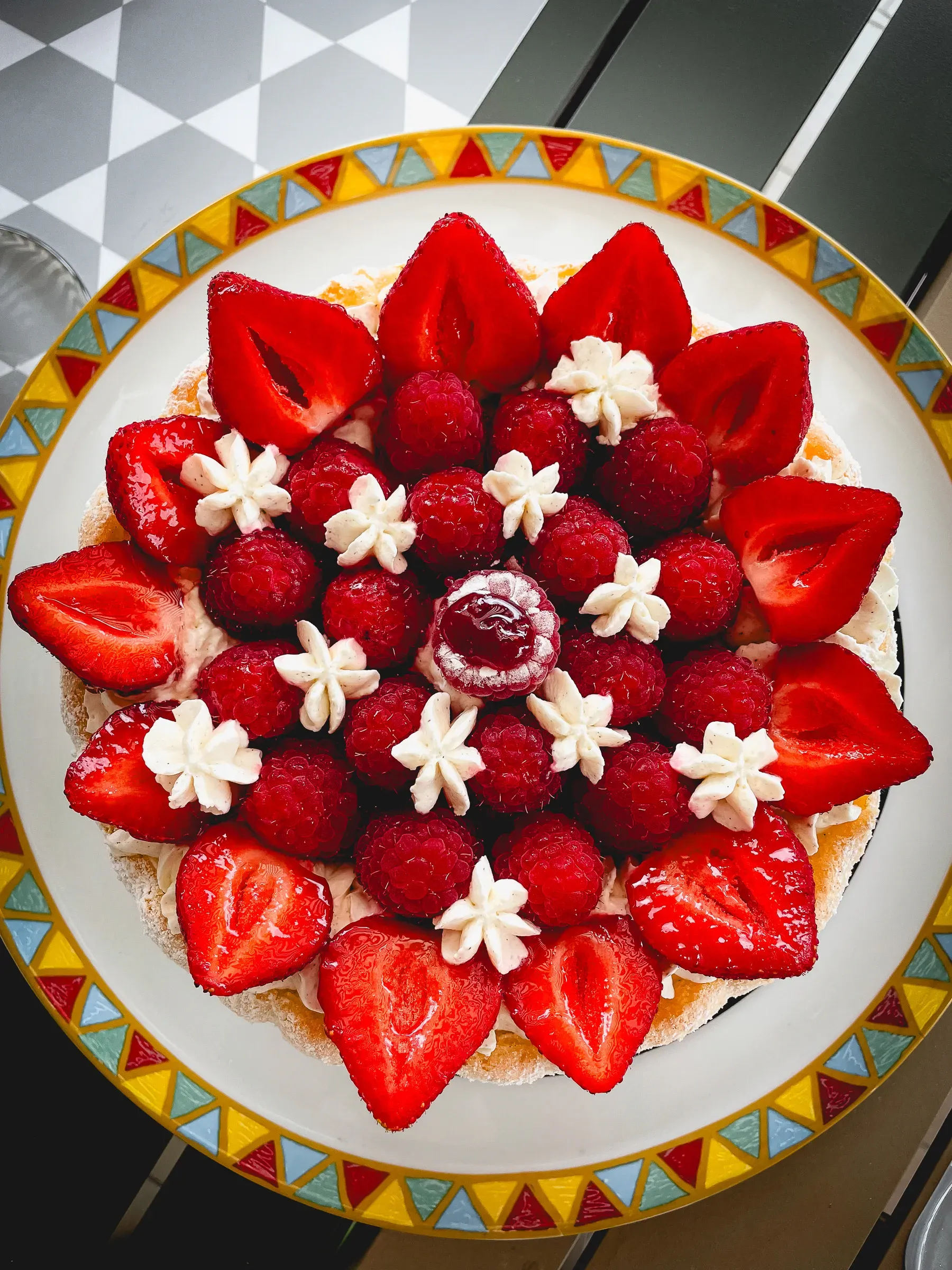 Strawberry and raspberry fraisier overhead on a decorative plate — strawberry halves and raspberries in a concentric pattern with cream rosettes, a red centre