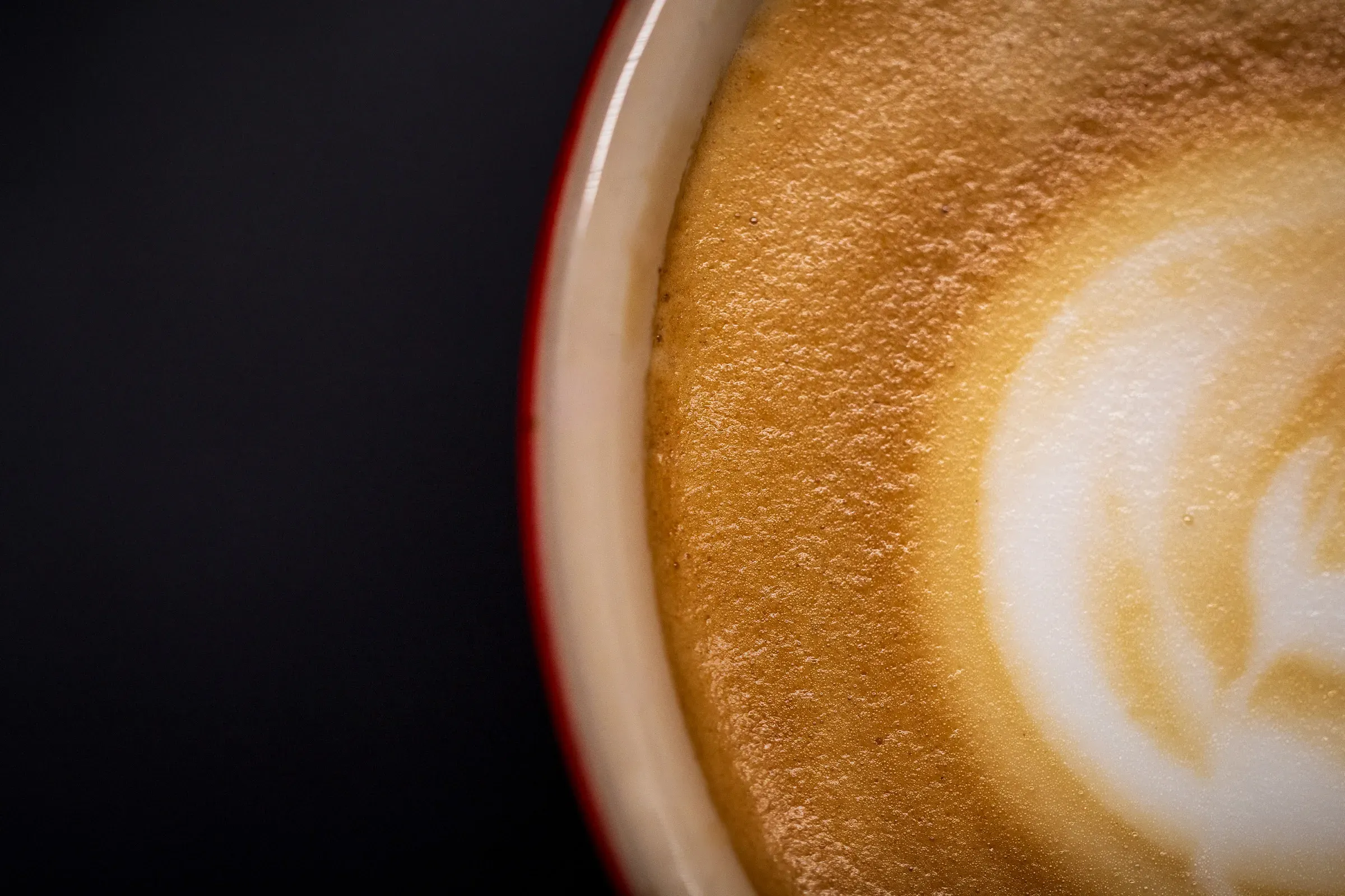 Latte in a red-rimmed cup, partial overhead frame — foam surface with latte art, dark background